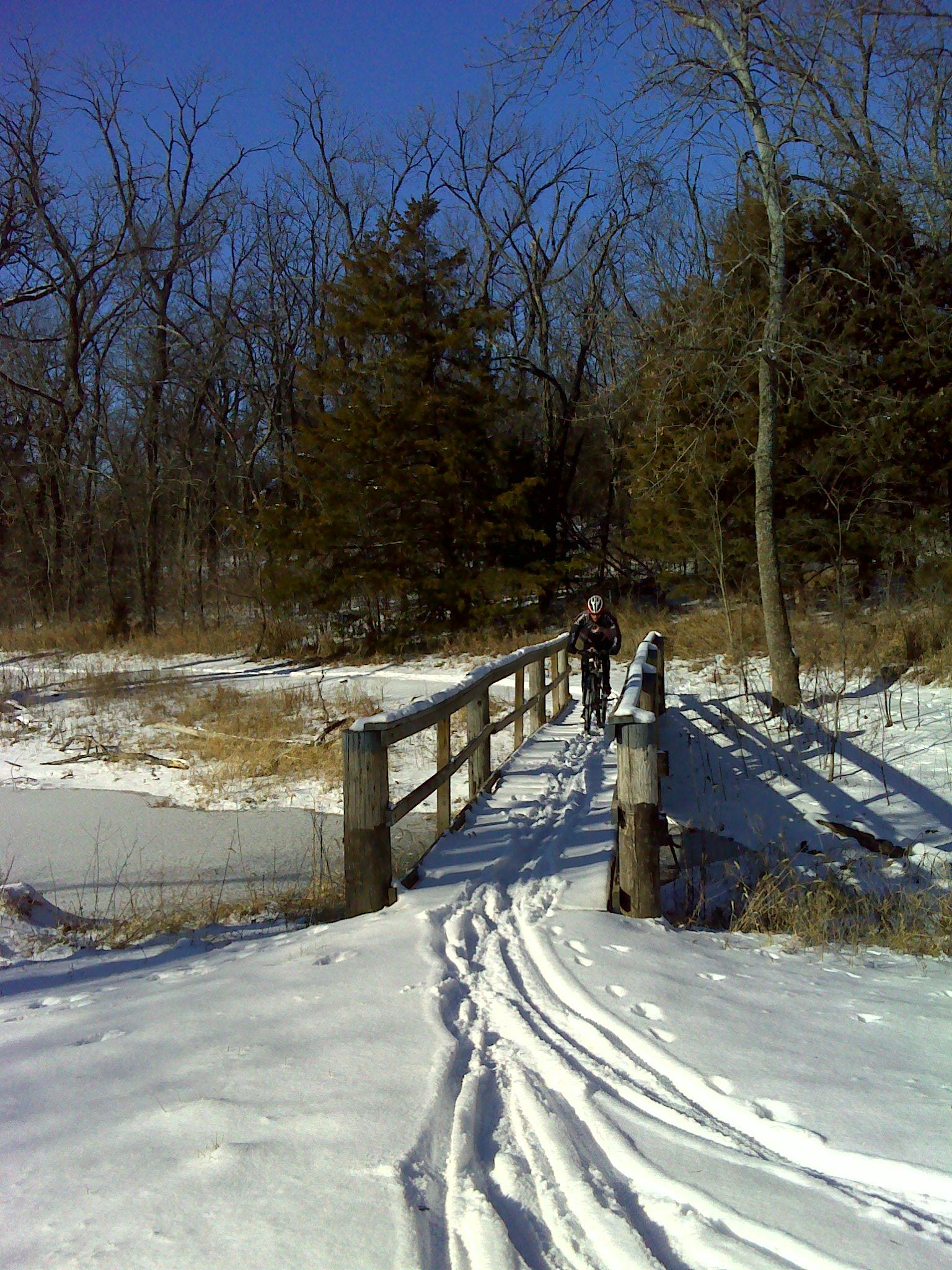 A person on a bicycle is crossing a wooden bridge surrounded by snow-covered ground and bare trees under a clear blue sky. Snow tracks lead up to the bridge, indicating recent activity in the winter landscape. Camp Alexander Trails mountain bike trail.