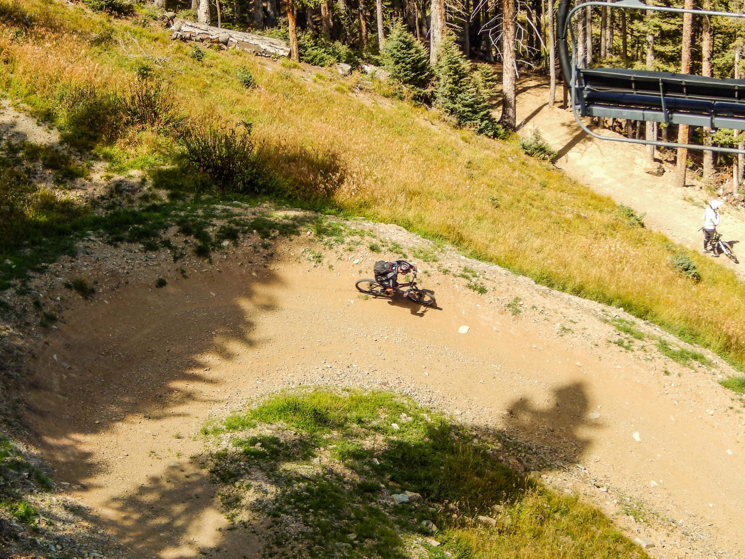 A cyclist riding through a dirt trail in a mountainous area, with surrounding trees and grass. A ski lift can be seen in the background, along with another cyclist in the distance. Taos Ski Valley mountain bike trail.
