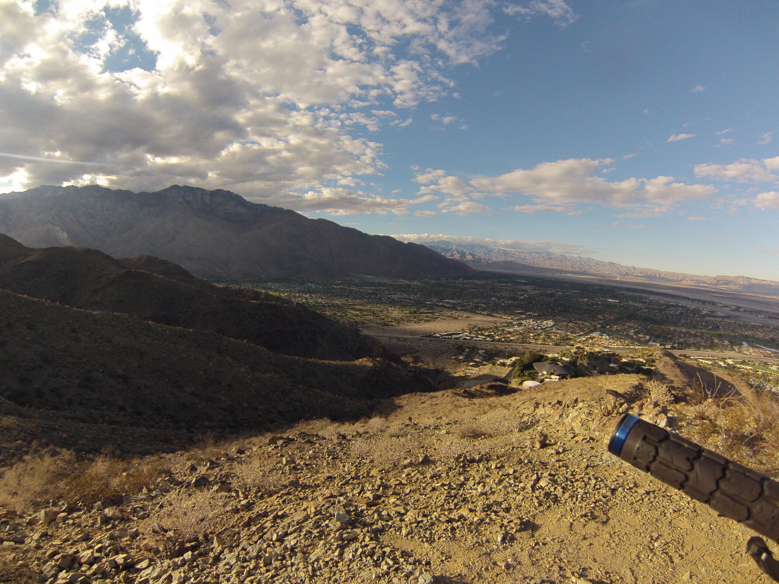 A panoramic view of a mountain landscape during the day, featuring rolling hills and a valley below. The sky is partly cloudy, with patches of blue visible among the clouds. In the foreground, a portion of a textured object, possibly a bike handle, is visible, hinting at an outdoor adventure. The background showcases the mountains and a nearby town nestled in the valley. The Goat Trails mountain bike trail.