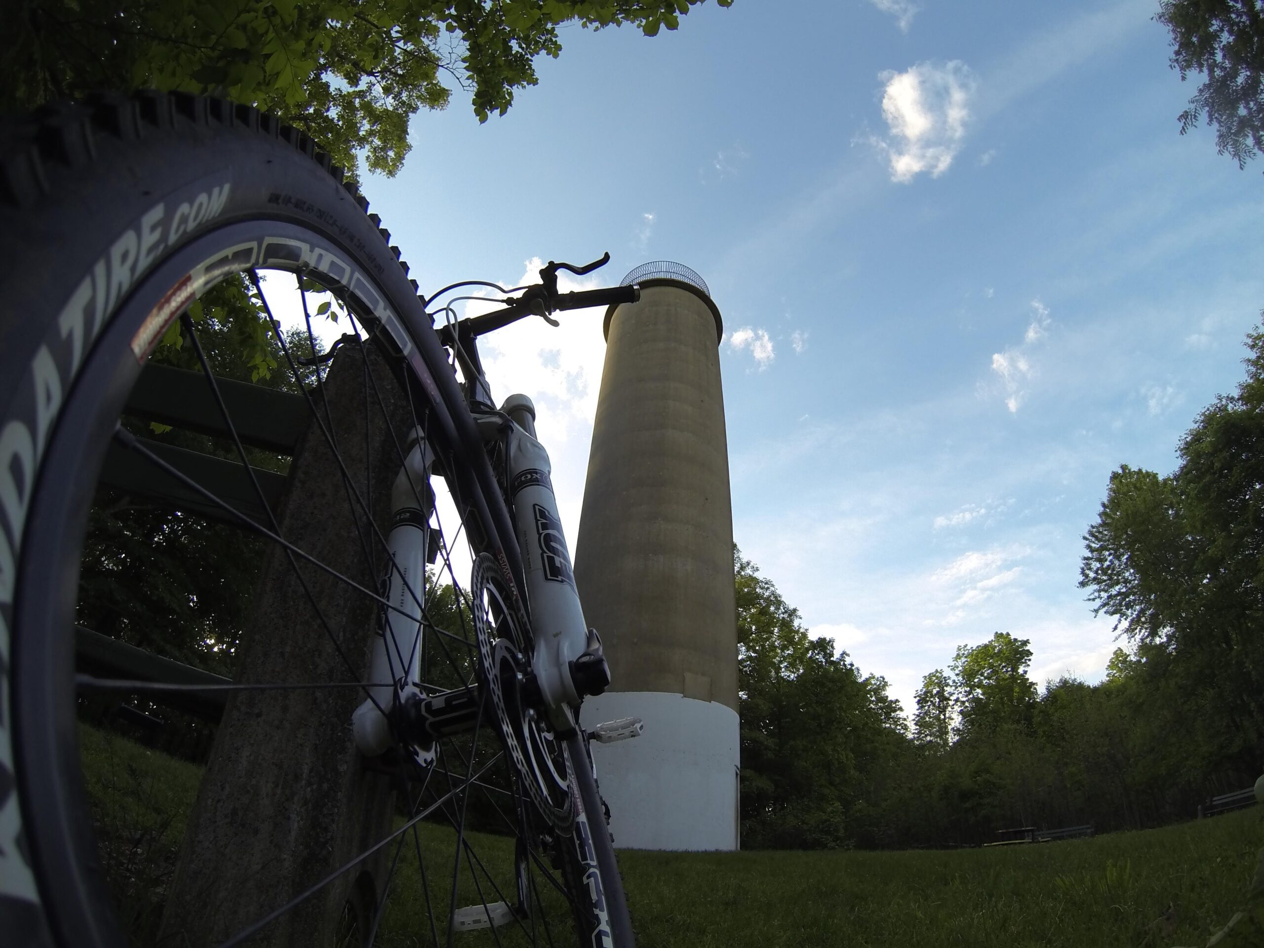 A low-angle view of a mountain bike leaning against a post, with a tall, cylindrical structure in the background. The scene is set under a clear blue sky with scattered clouds, surrounded by greenery and trees. Governor Dick mountain bike trail.