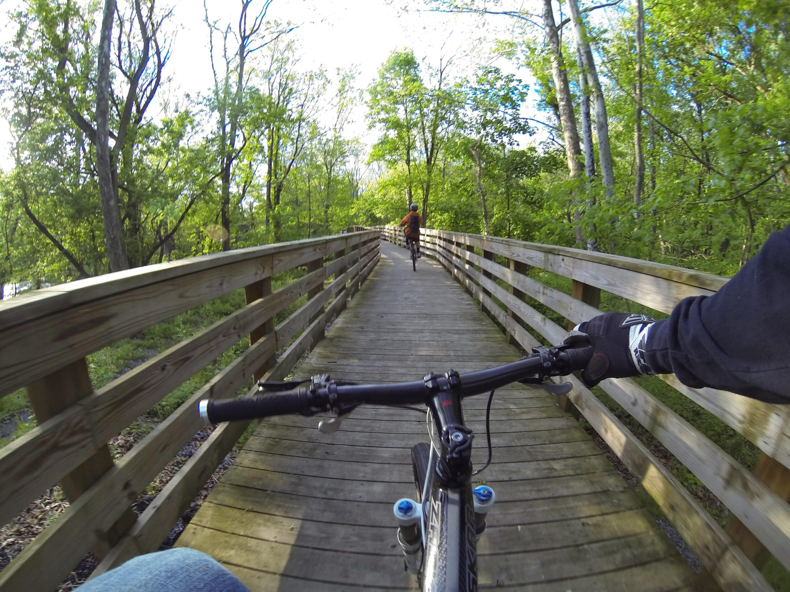 A scenic view of a bike path through a wooded area, showcasing a wooden bridge that cyclists are riding on. The image captures the handlebars of one bike in the foreground, while another cyclist is seen in the distance, surrounded by lush greenery and sunlight filtering through the trees. Quittie Creek Nature Park mountain bike trail.