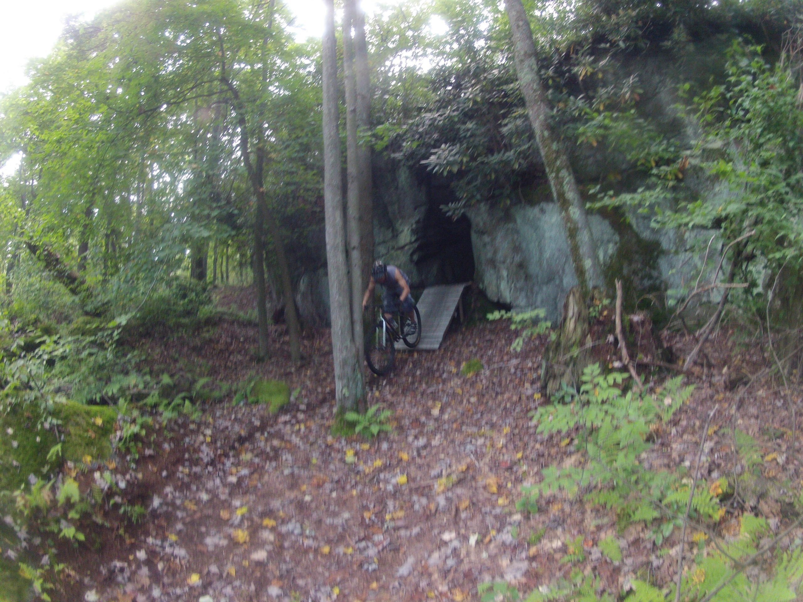 A mountain biker navigating through a forested area with a rocky outcrop and a small wooden ramp, surrounded by trees and fallen leaves. Big Bear Lake Trail Center mountain bike trail.
