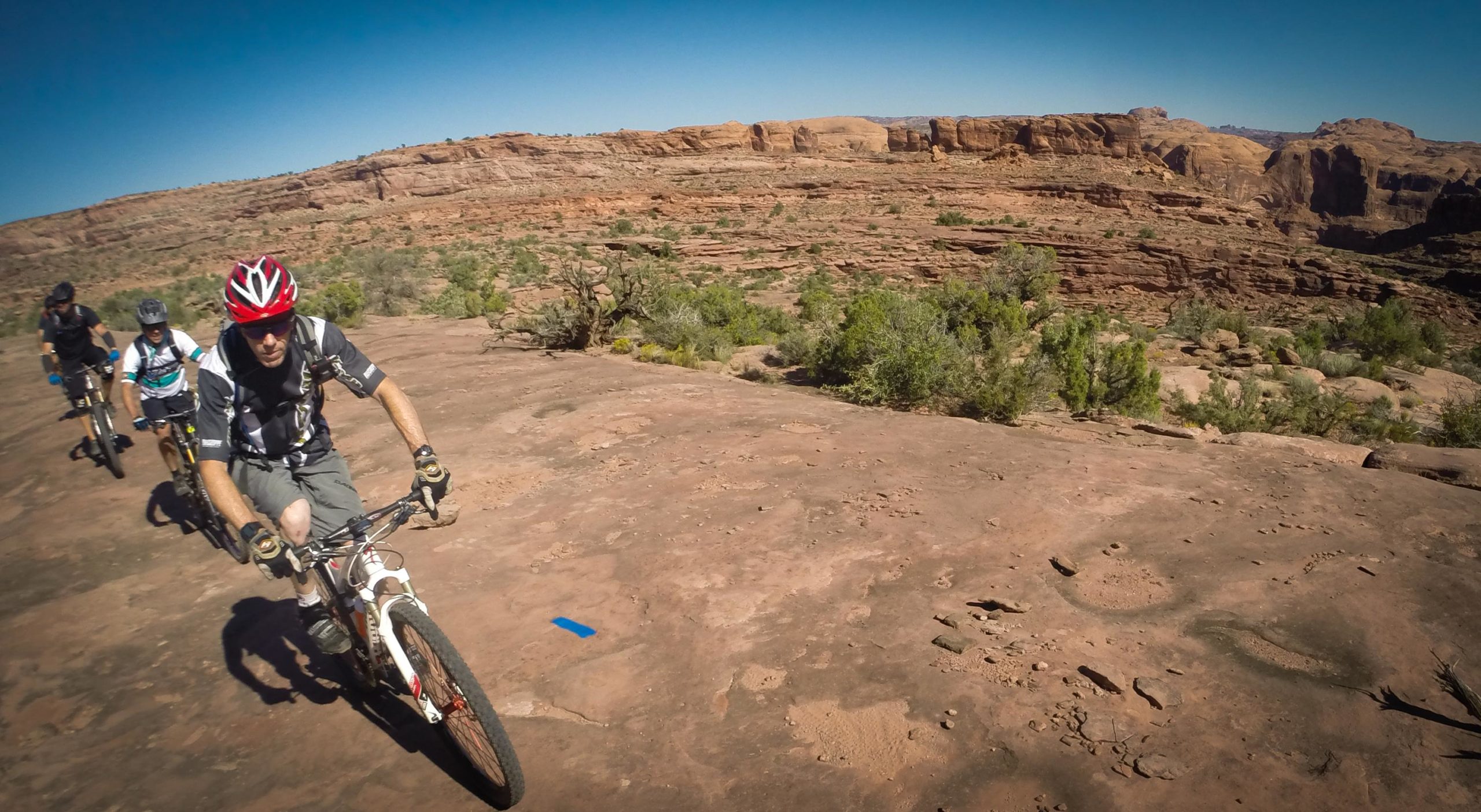 A group of four mountain bikers riding along a rocky trail in a desert landscape, with red rock formations and sparse vegetation in the background under a clear blue sky. Hymasa mountain bike trail.