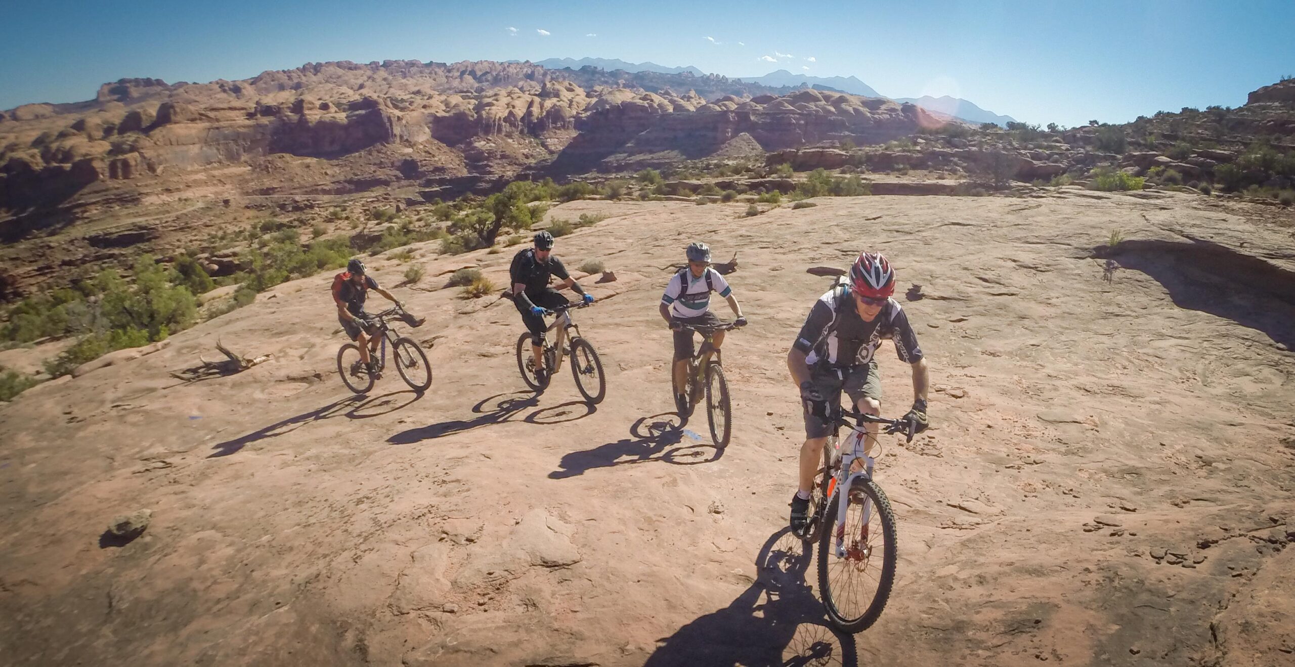 Four mountain bikers riding on rocky terrain with a desert landscape in the background, featuring rugged cliffs and distant mountains under a clear blue sky. Hymasa mountain bike trail.