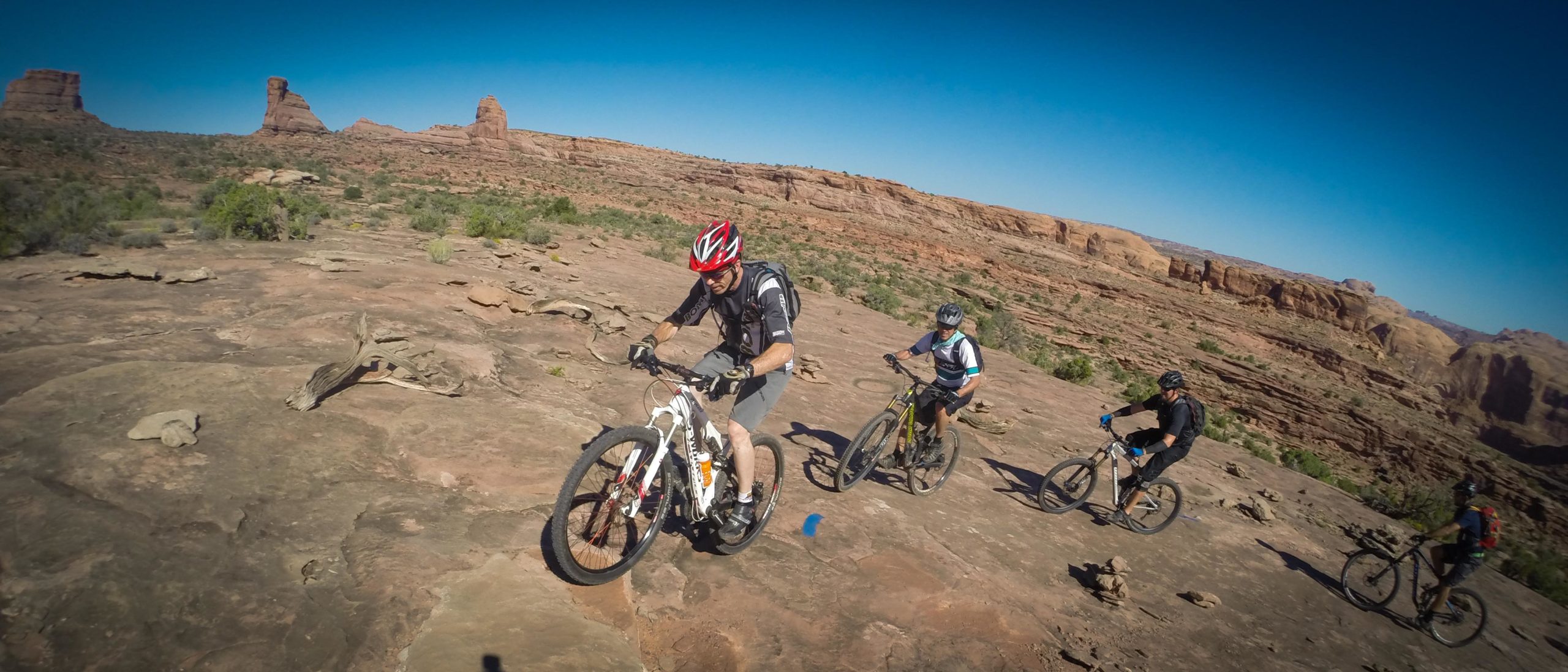 A group of four mountain bikers navigating a rocky terrain under a clear blue sky. The riders are wearing helmets and riding on a trail surrounded by desert landscapes, with distant rock formations in the background. Hymasa mountain bike trail.