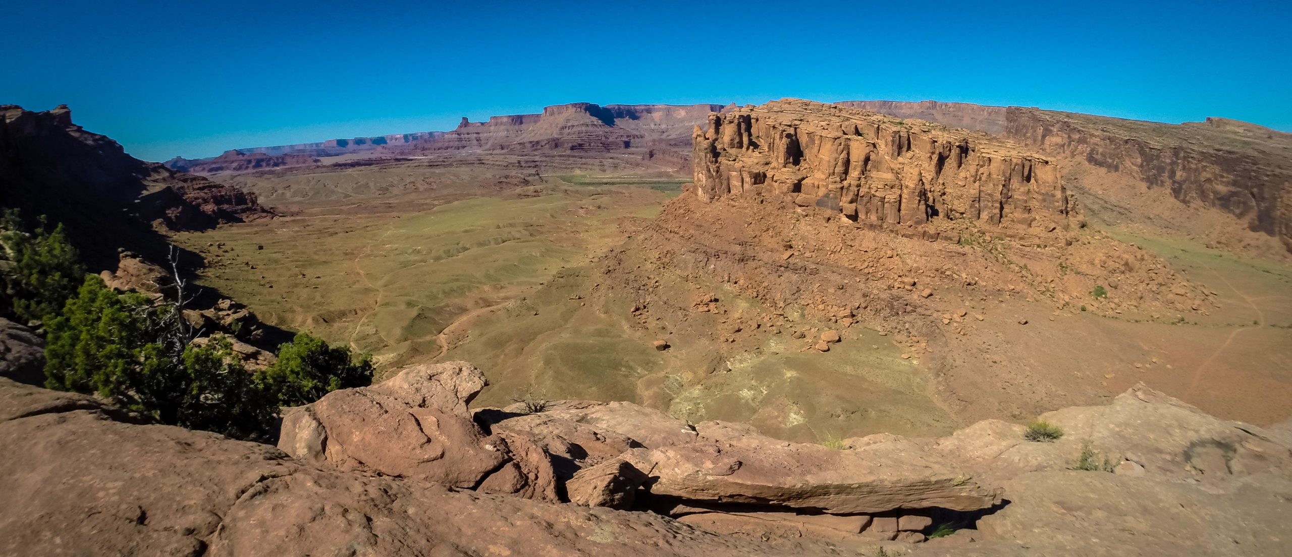 A panoramic view of a rugged desert landscape featuring towering rock formations and vast stretches of green terrain under a clear blue sky. The scene showcases the natural beauty and geological features characteristic of arid environments. Hymasa mountain bike trail.