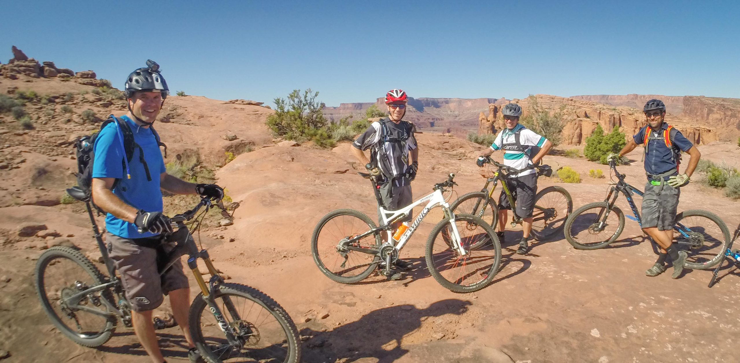 A group of four mountain bikers posing for a photo on a rocky landscape, surrounded by desert vegetation and blue sky. Each rider is dressed in cycling gear and standing next to their bikes, smiling at the camera. The backdrop features rock formations typical of a desert environment. Hymasa mountain bike trail.