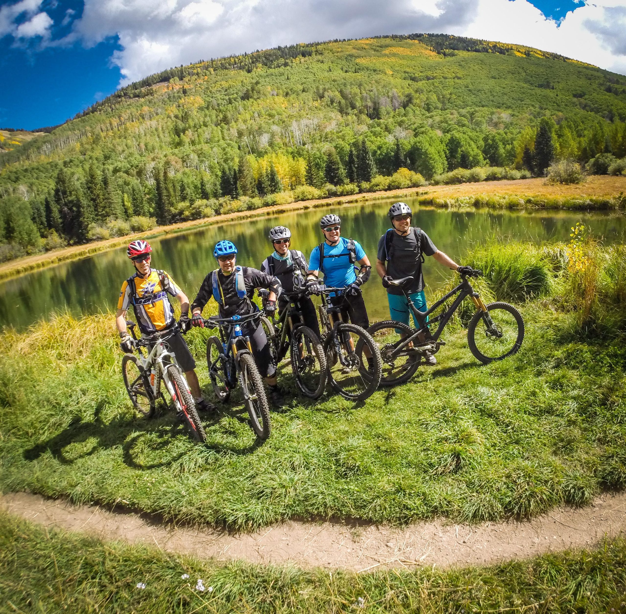 Five mountain bikers pose by a serene pond surrounded by lush green trees and rolling hills. They are wearing helmets and cycling gear, standing with their bikes on a grassy patch near a dirt path. The sky is partly cloudy, indicating a bright, sunny day. The Whole Enchilada mountain bike trail.