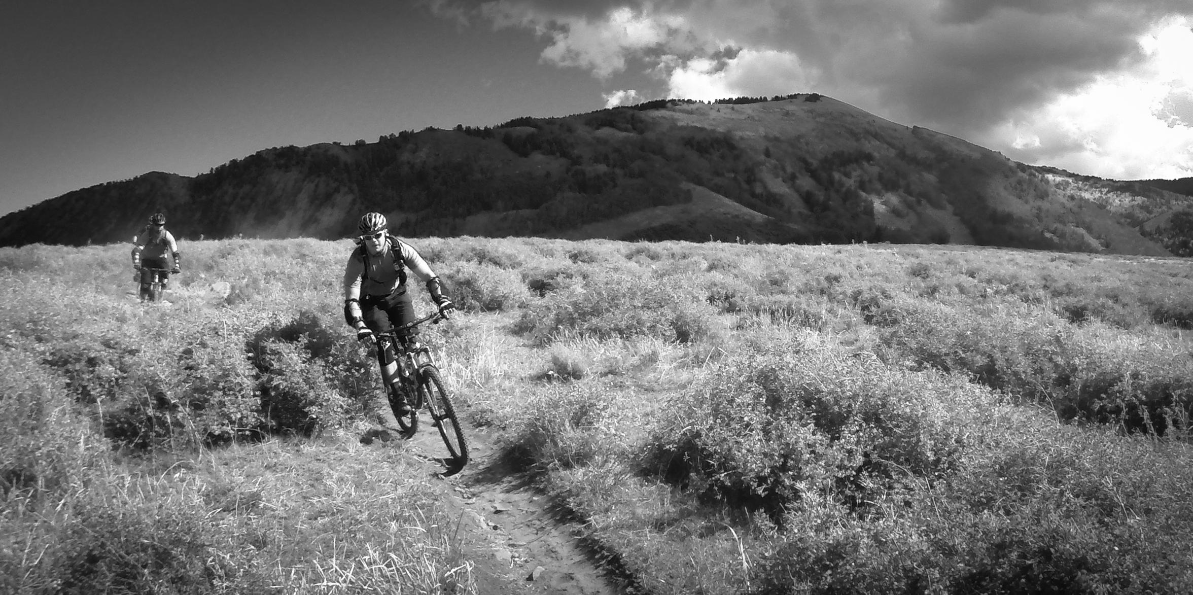 Two mountain bikers ride along a dirt trail through a grassy landscape, with a mountainous backdrop under a cloudy sky. The image is in black and white, highlighting the contours of the terrain and the bikers' movements. The Whole Enchilada mountain bike trail.