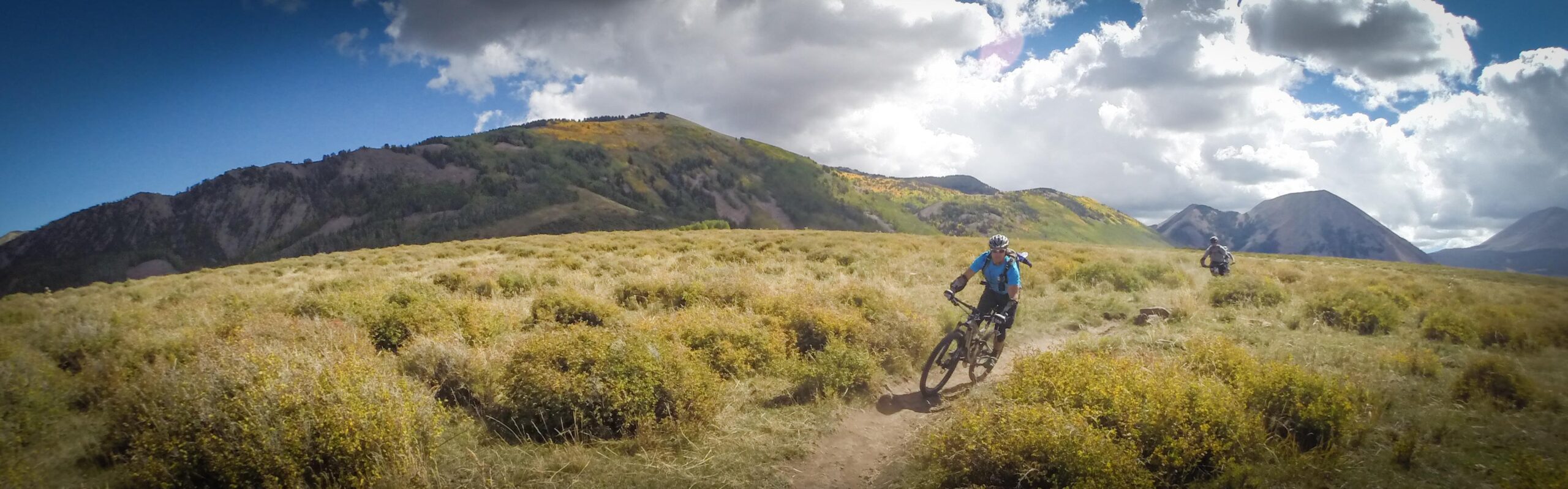 Two mountain bikers ride along a dirt trail through a grassy landscape, surrounded by rolling hills and mountains under a partly cloudy sky. The vibrant fall colors of the trees in the background add warmth to the scene. The Whole Enchilada mountain bike trail.