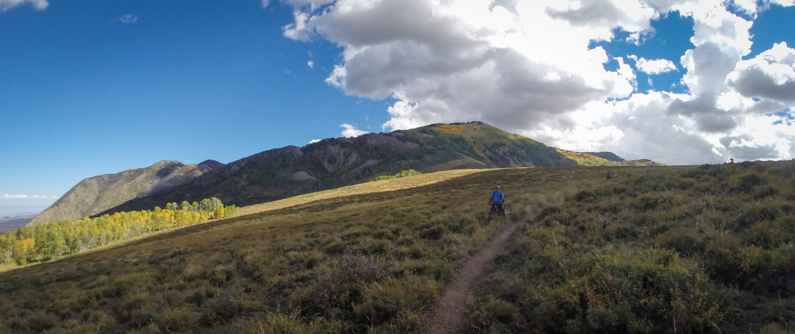 A panoramic view of a mountainous landscape under a blue sky with scattered clouds. A hiker in blue is riding a bike along a dirt trail winding through grassy terrain, with patches of trees changing colors in the foreground. Another person can be seen in the distance, also biking. The scene captures the beauty of nature and outdoor adventure. The Whole Enchilada mountain bike trail.