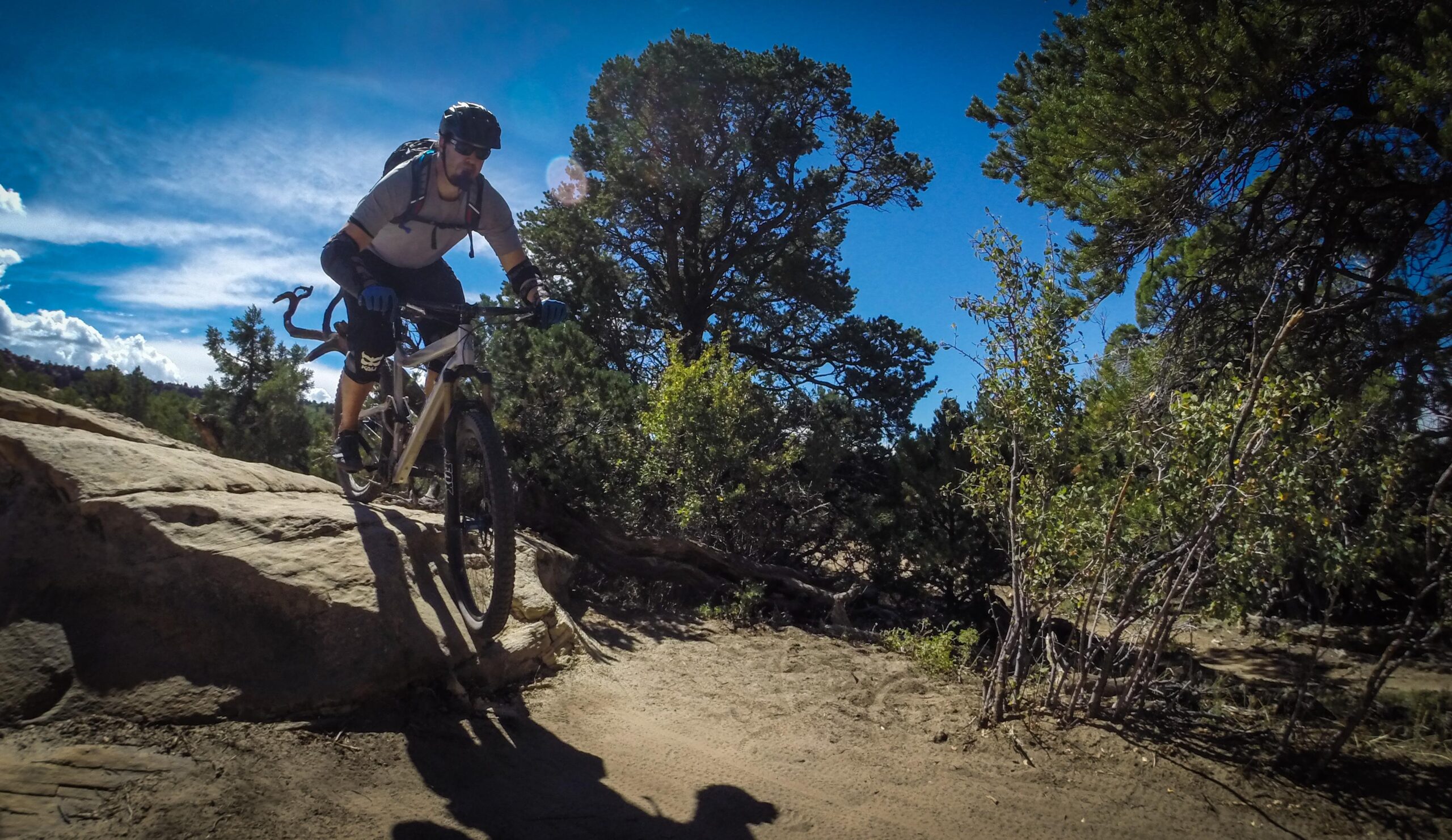 A mountain biker riding over a rocky terrain with trees in the background under a clear blue sky. The bike has thick tires, and the rider is wearing a helmet and gloves, focused on navigating the trail. The Whole Enchilada mountain bike trail.