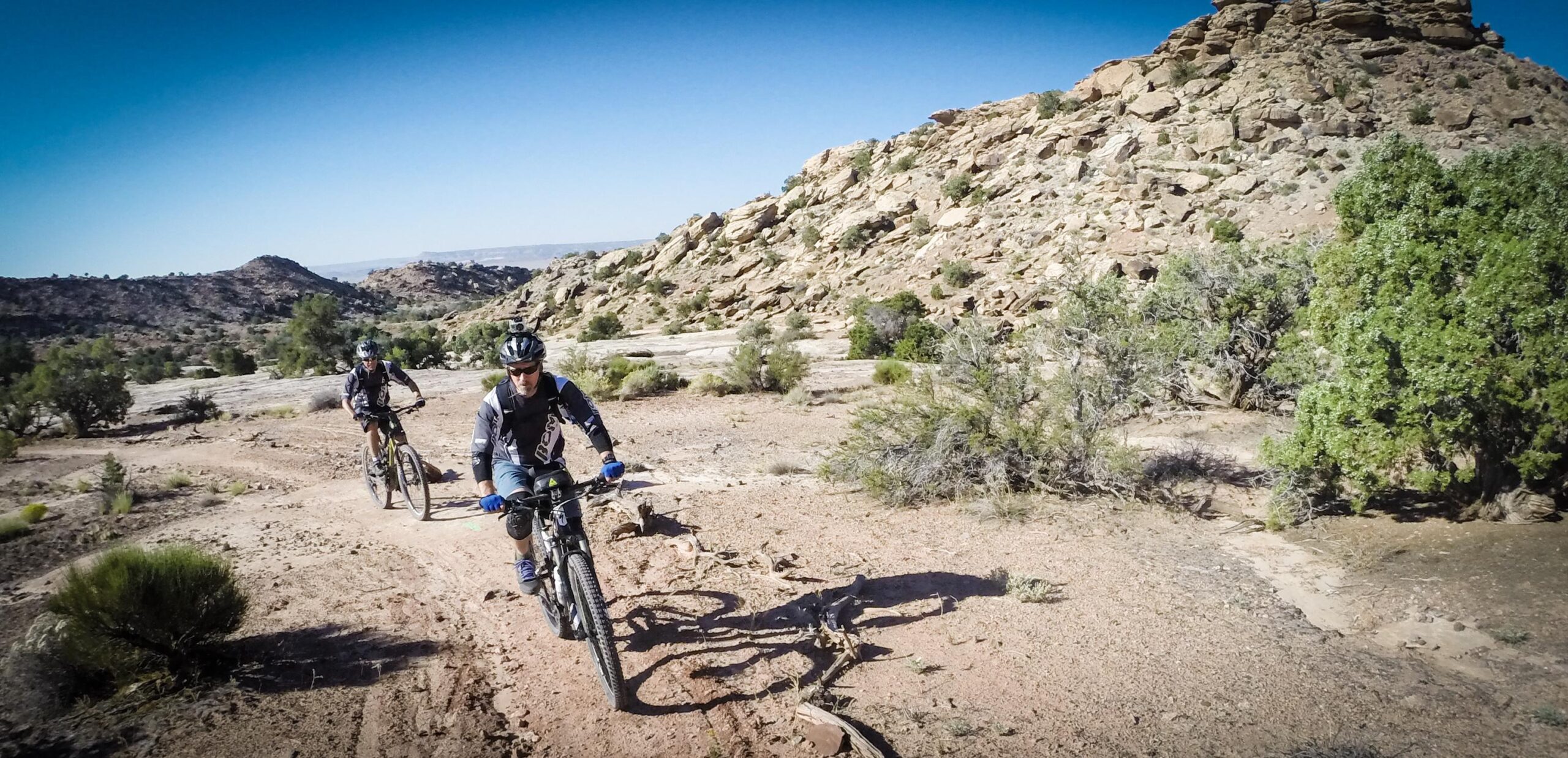 Two mountain bikers riding on a dirt trail in a rocky, desert landscape. The background features rugged terrain with hills and sparse vegetation under a clear blue sky. Klondike Bluffs mountain bike trail.
