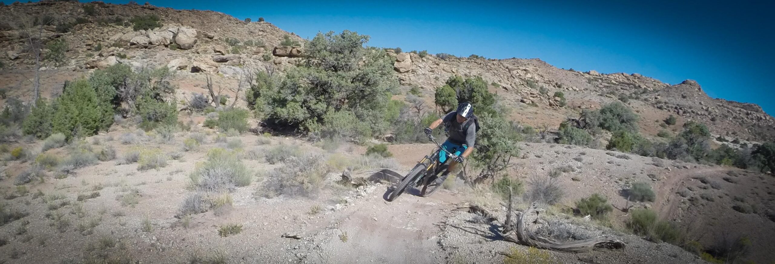 A mountain biker navigating a rocky trail in a desert landscape, performing a jump with dust and debris kicking up behind. The scene shows rugged terrain with sparse vegetation and steep hills in the background under a clear blue sky. Klondike Bluffs mountain bike trail.