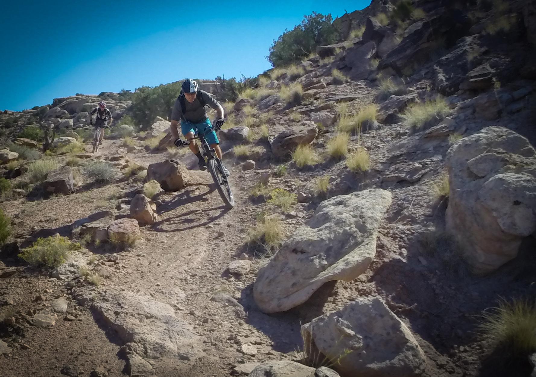 Mountain bikers maneuvering through a rocky trail in a desert landscape under a clear blue sky. The foreground features one rider leaning into a turn, while another biker is visible in the background. Surrounding vegetation includes sparse grass and shrubs amid the rugged terrain. Klondike Bluffs mountain bike trail.