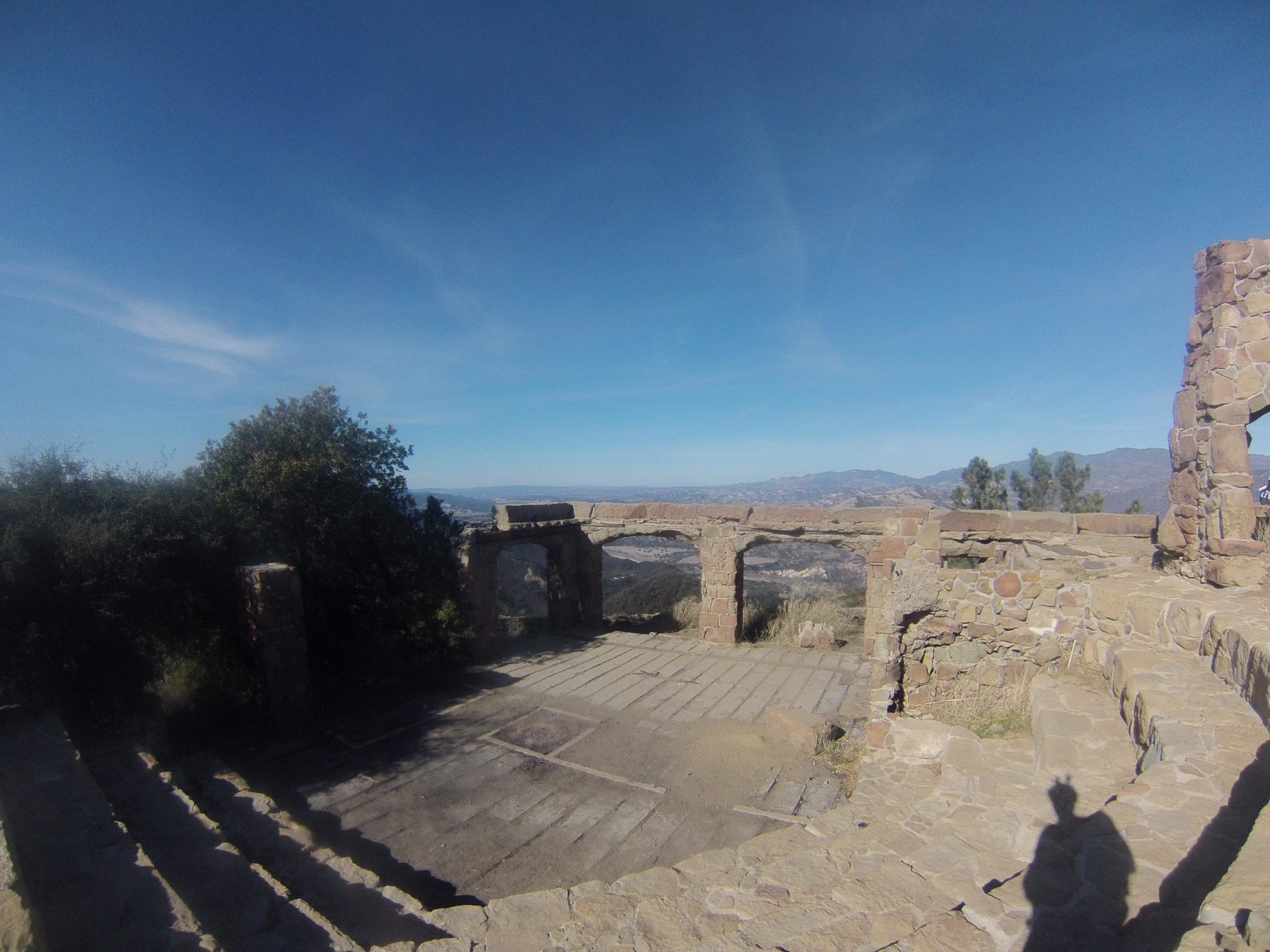 An expansive view of ancient stone ruins against a clear blue sky. The image features remnants of a structure made from large stones, with steps leading down and a backdrop of distant mountains and trees. The scene captures the serene atmosphere of the site, highlighting the blending of nature and history. Snyder Trail / Knapps Castle mountain bike trail.