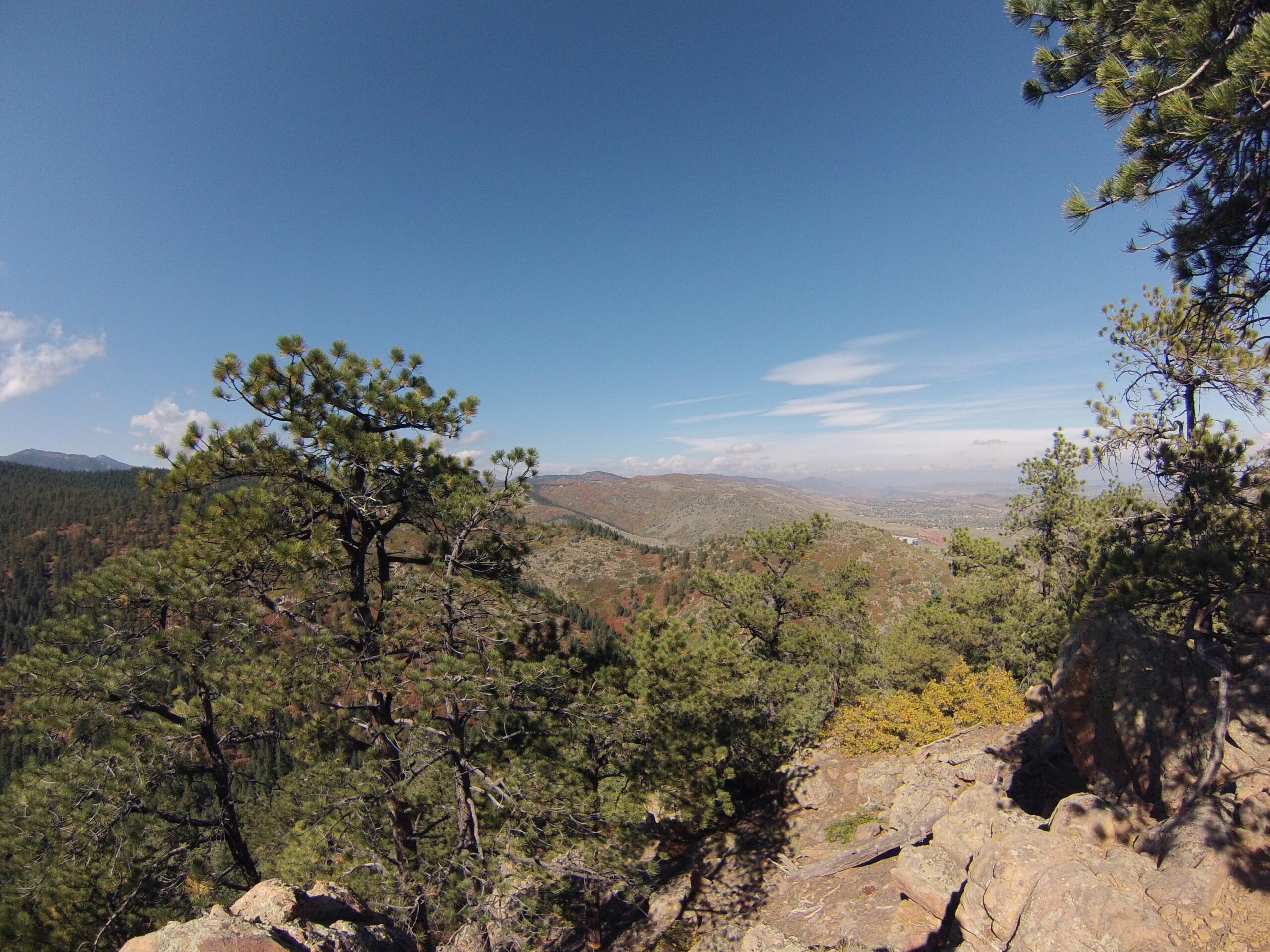 A panoramic view of a mountainous landscape featuring dense green forests, scattered rocky outcrops, and a clear blue sky with a few clouds. In the foreground, there are several pine trees and boulders, while the background reveals rolling hills and distant mountains. Deer Creek Canyon mountain bike trail.