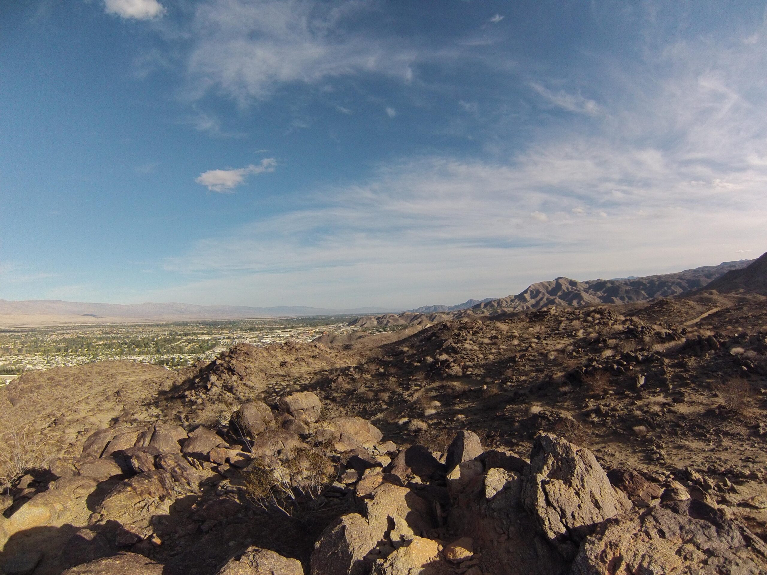 A panoramic view of a desert landscape featuring rocky terrain, distant mountains, and a clear blue sky with scattered clouds. A small town is visible below amidst the arid environment. The Goat Trails mountain bike trail.