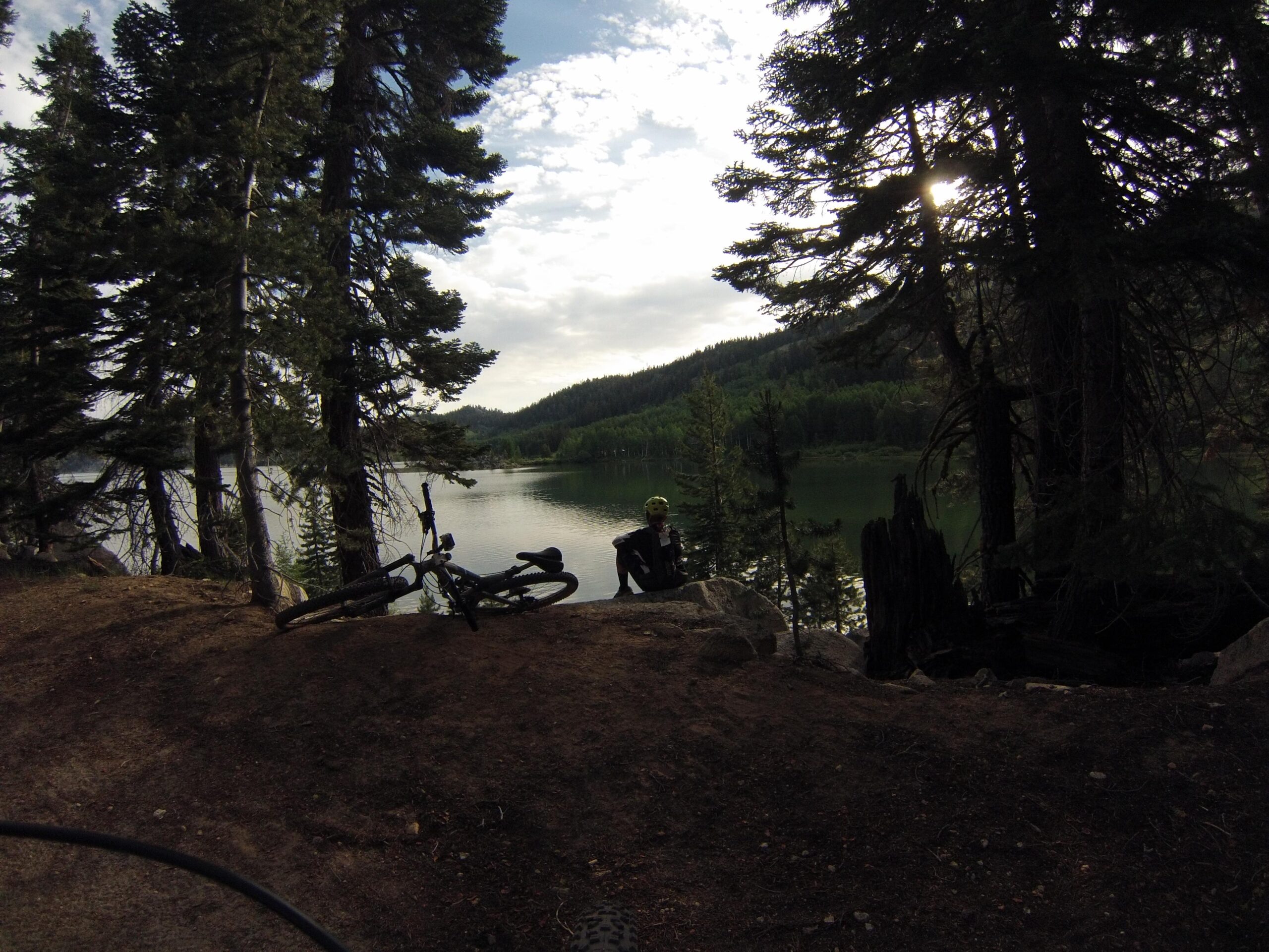 A peaceful lakeside scene surrounded by tall trees, with a bicycle resting on the ground. In the foreground, a person sits on a rock near the water, enjoying the serene atmosphere and beautiful natural landscape. The sun peeks through the trees, casting gentle light on the tranquil scene. Flume Trail mountain bike trail.