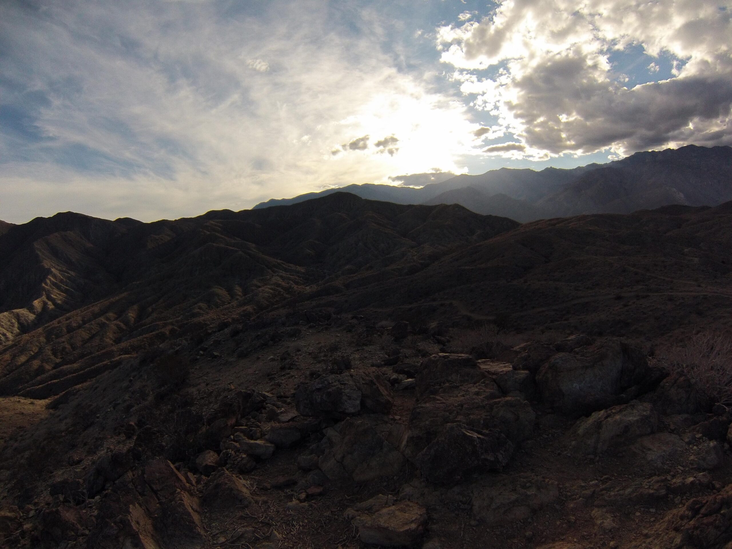 A mountainous landscape under a cloudy sky, with rolling hills and rocky terrain, illuminated by sunlight peeking through the clouds. The scene captures a sense of tranquility and the natural beauty of the outdoors. The Goat Trails mountain bike trail.