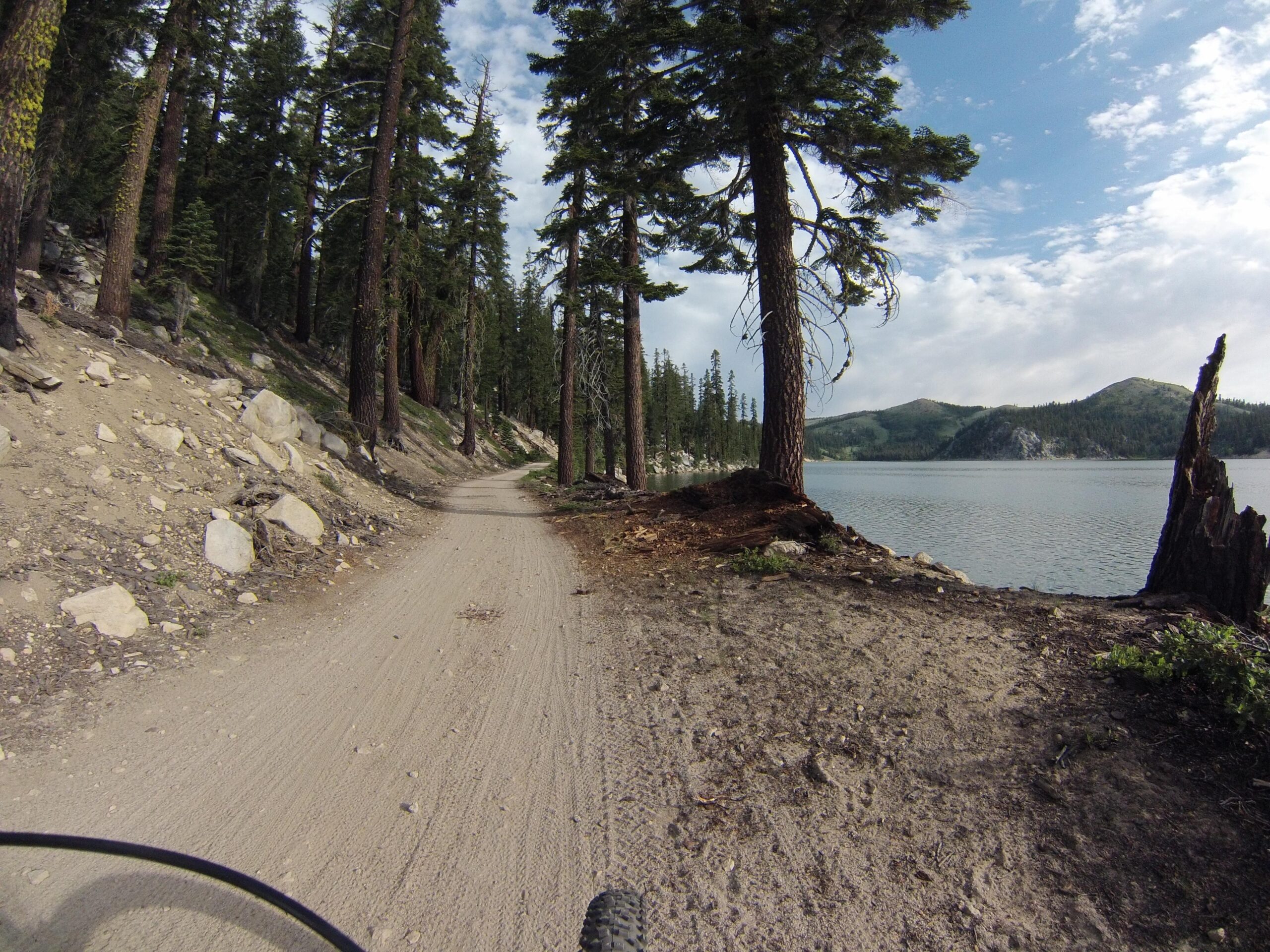 A scenic dirt path winds through a forest of tall pine trees, with large rocks scattered along the trail. To the right, a calm lake reflects the surrounding mountains under a partly cloudy sky. The image captures a tranquil outdoor atmosphere, ideal for biking or hiking. Flume Trail mountain bike trail.