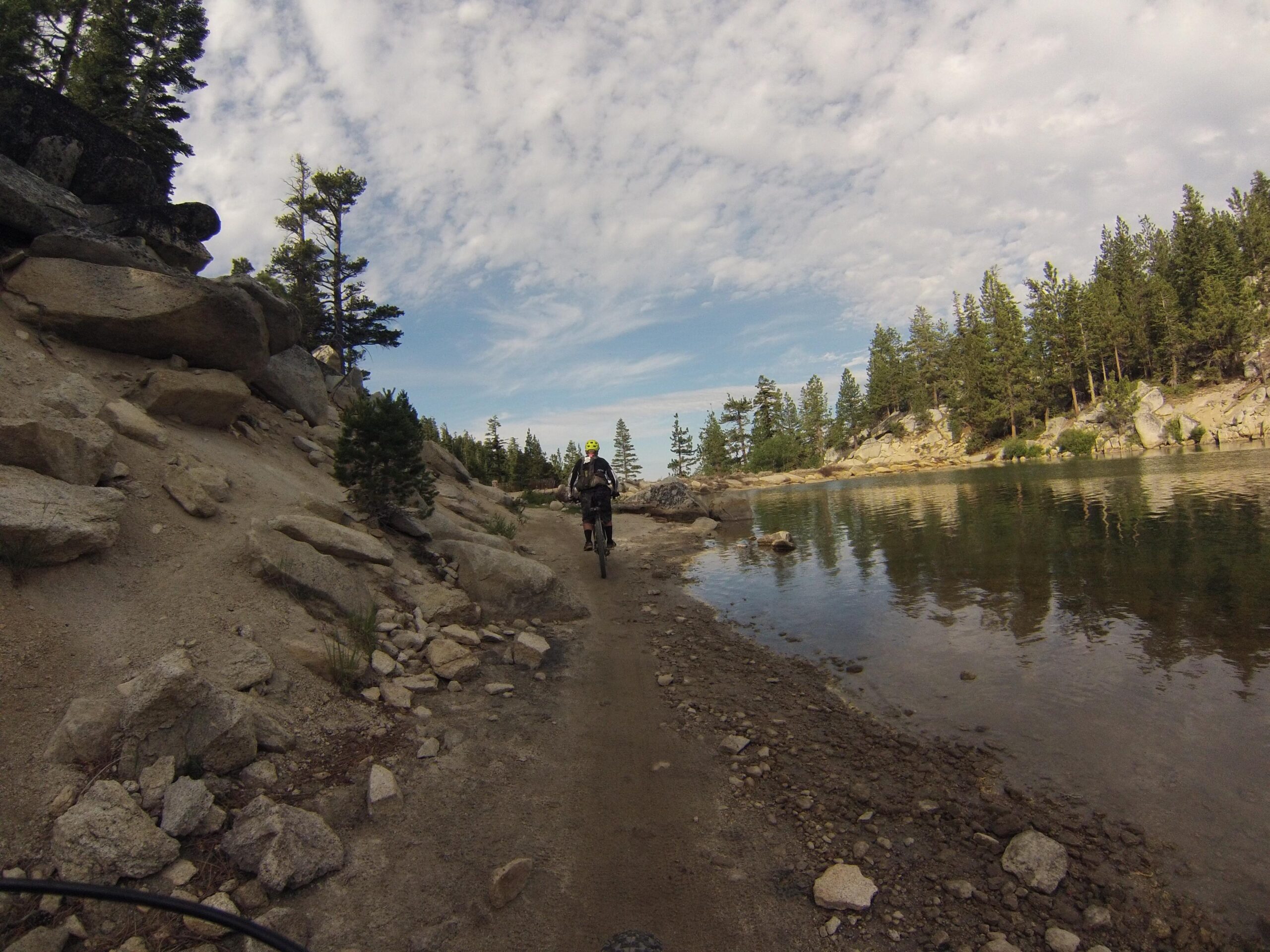 A person riding a mountain bike on a rocky path beside a calm body of water, surrounded by trees and large boulders under a partly cloudy sky. Flume Trail mountain bike trail.