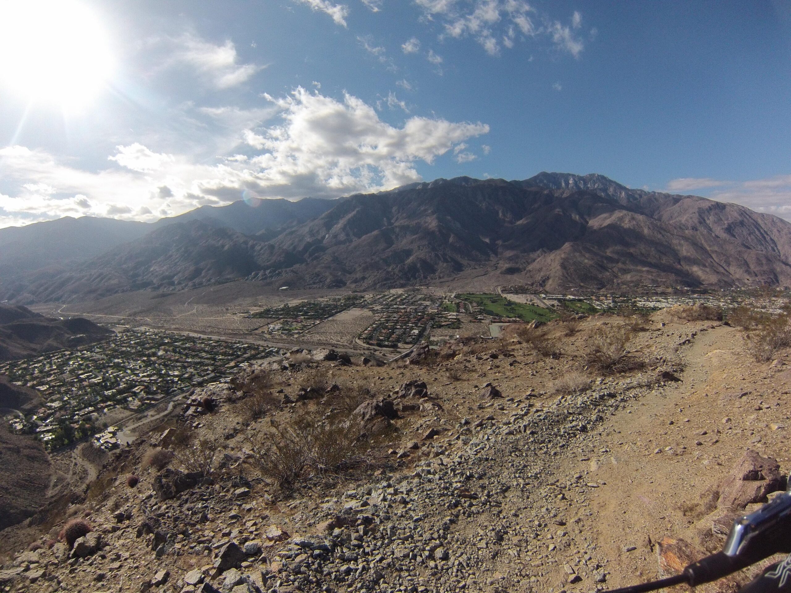 A panoramic view from a mountainside overlooking a desert valley, with homes and green spaces visible below. The landscape features rugged mountains in the background, partly cloudy skies, and sunlight illuminating the scene. The Goat Trails mountain bike trail.