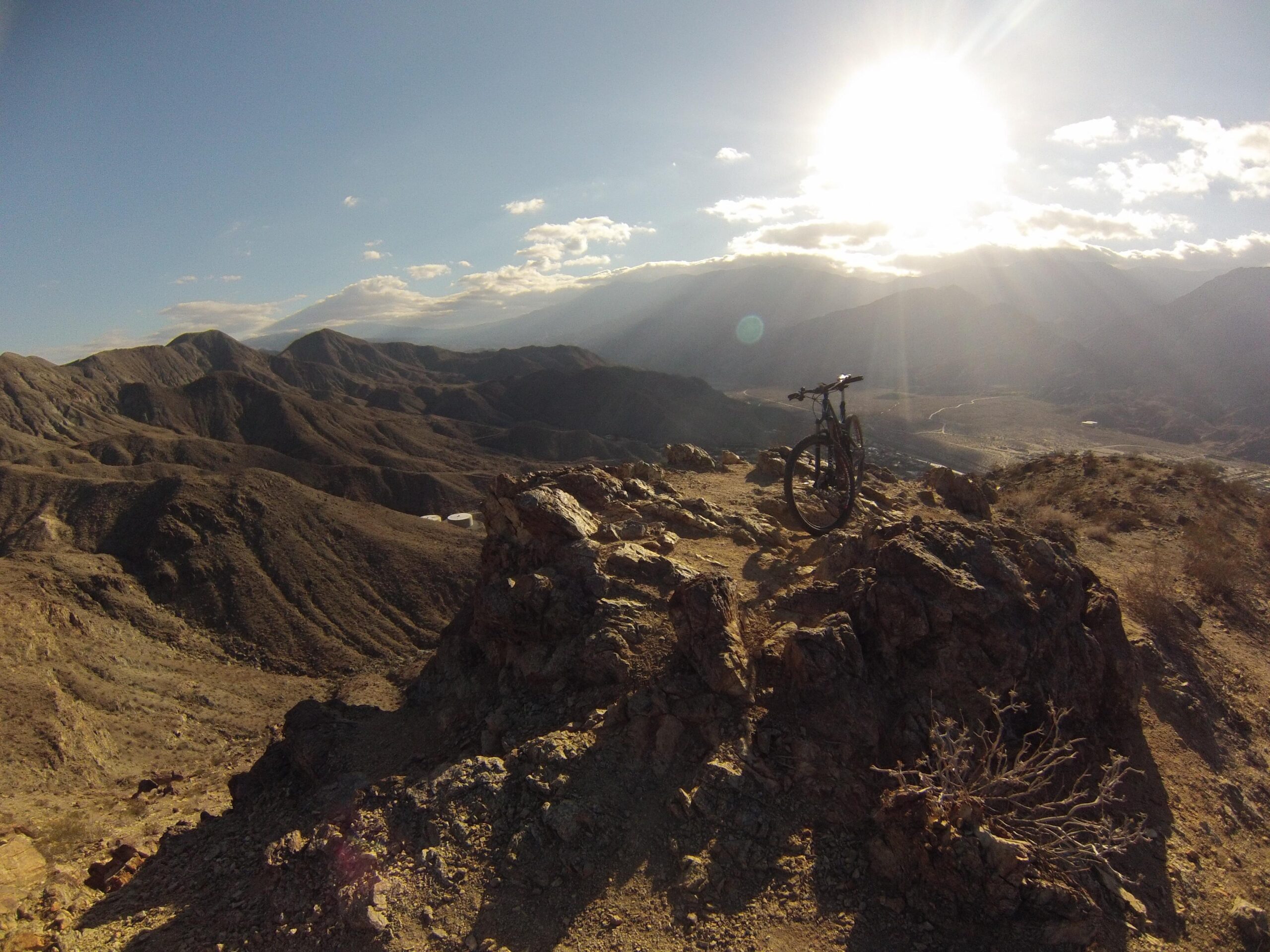 A mountain bike resting on a rocky outcrop overlooking a scenic mountainous landscape under a bright sun. The terrain is rugged and dry, with hills and valleys stretching into the distance. The Goat Trails mountain bike trail.