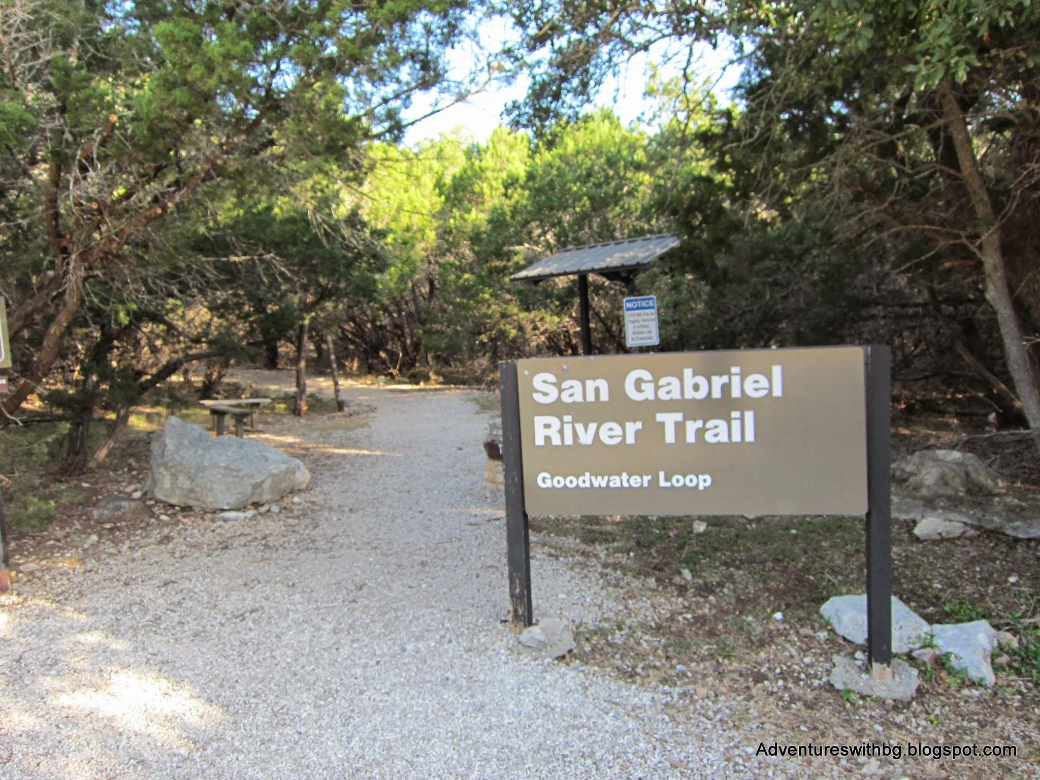 A sign at the entrance of the San Gabriel River Trail, indicating the Goodwater Loop. The trail is surrounded by lush greenery, with a gravel path leading into the woods. There is also a small picnic table visible nearby. Goodwater Trail mountain bike trail.