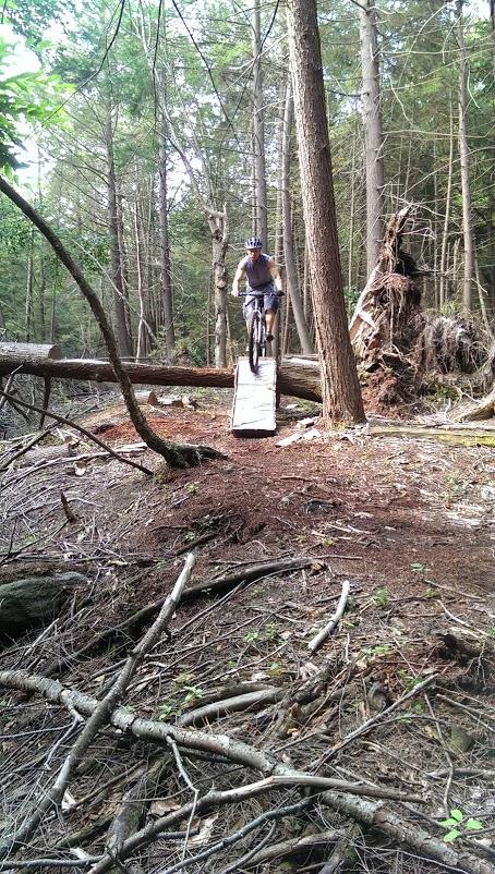 A person on a mountain bike riding over a narrow wooden bridge in a dense forest setting, surrounded by trees and fallen branches. Big Bear Lake Trail Center mountain bike trail.