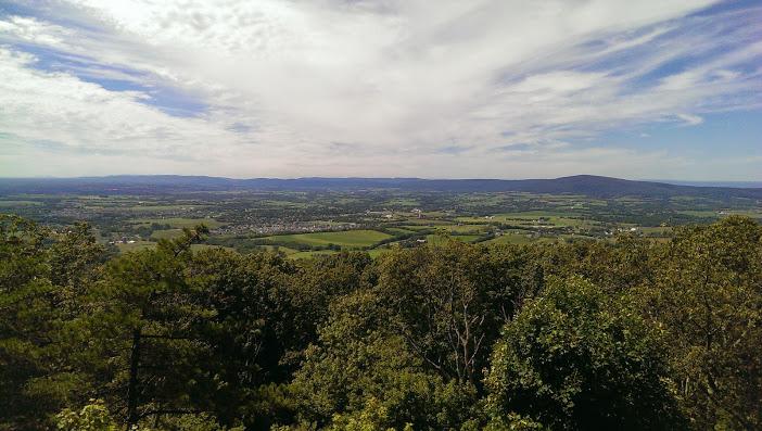 A panoramic view of lush green valleys and distant mountains under a partly cloudy sky, with trees in the foreground. Gambrill State Park mountain bike trail.
