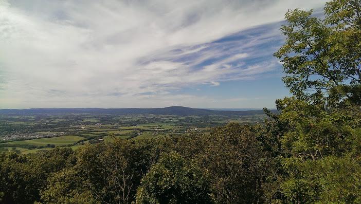 A panoramic view of rolling green hills and valleys under a partly cloudy sky, with trees in the foreground. The landscape features distant mountains and a meandering river, showcasing a serene natural setting. Gambrill State Park mountain bike trail.