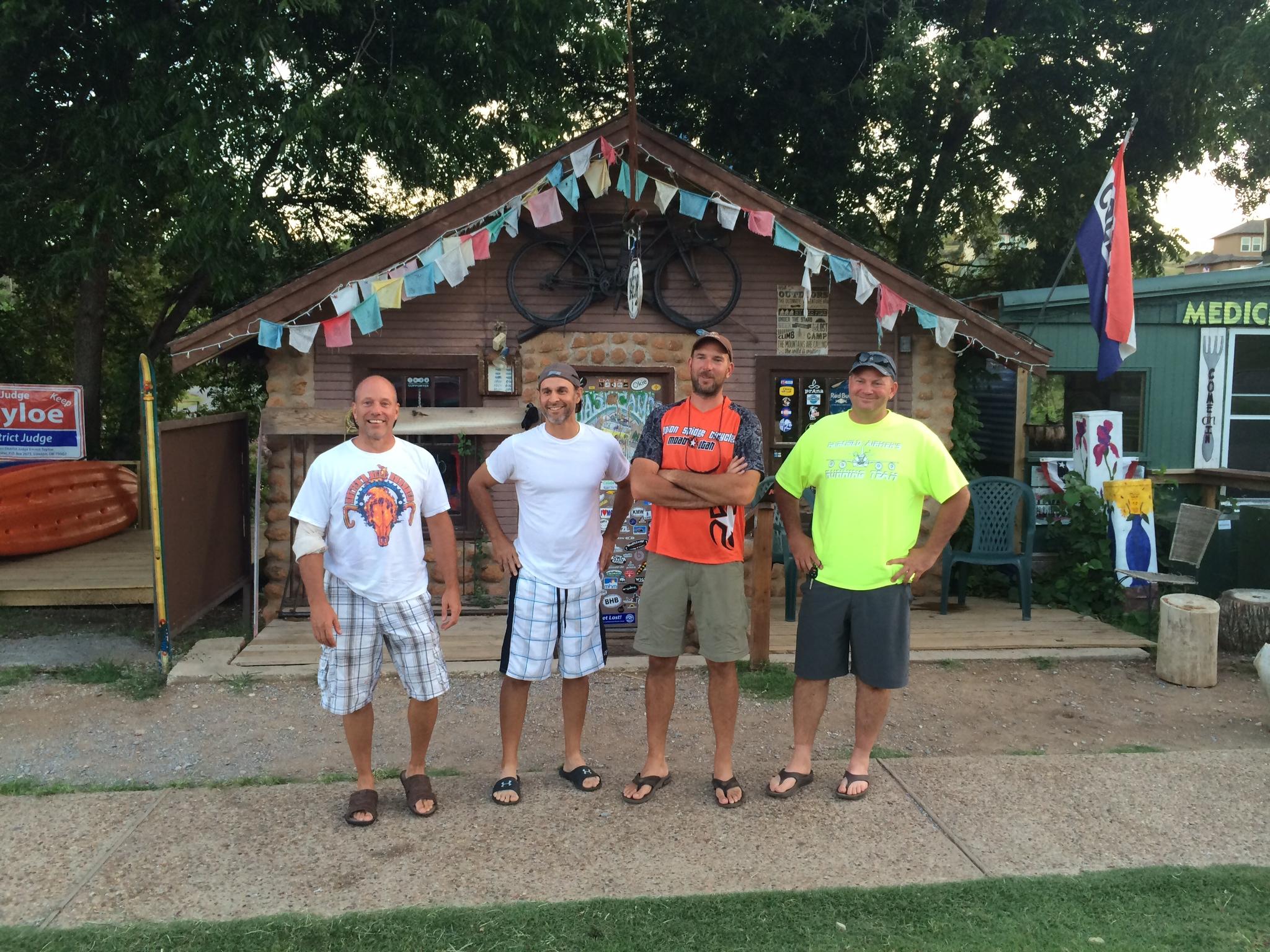 Four men standing in front of a quaint cabin-like building, smiling and posing. The building features decorative elements like prayer flags and a bicycle mounted on the wall. They are dressed in casual summer attire, with shorts and flip-flops. In the background, there are kayaks and signs visible, hinting at an outdoor recreational theme. The setting appears relaxed and inviting, surrounded by greenery. Lake Lawtonka Trails mountain bike trail.