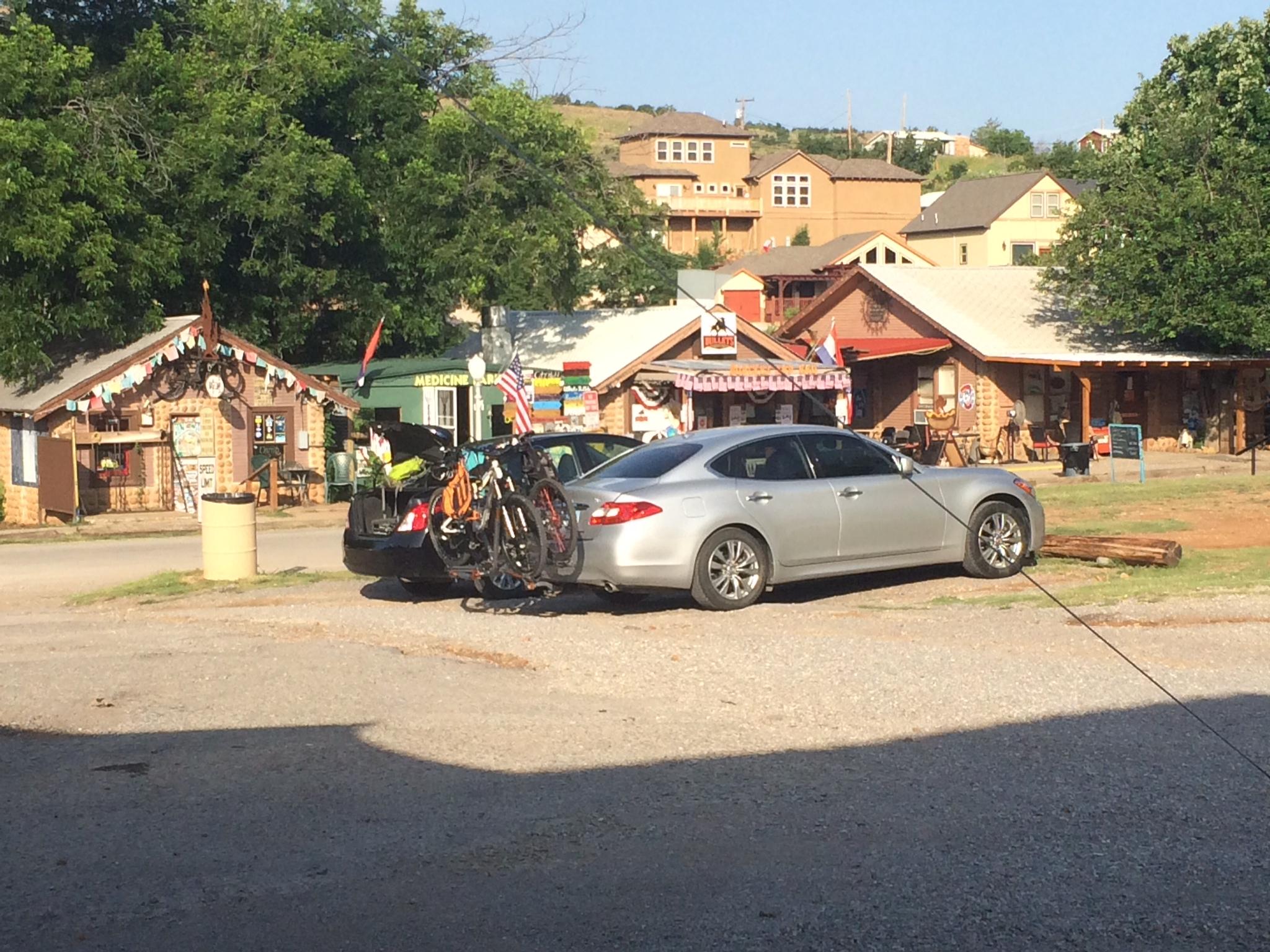 A silver car with bicycles mounted on a rack parked in front of rustic shops with decorative signage. The scene includes green trees and a clear blue sky, suggesting a vibrant outdoor setting. Lake Lawtonka Trails mountain bike trail.