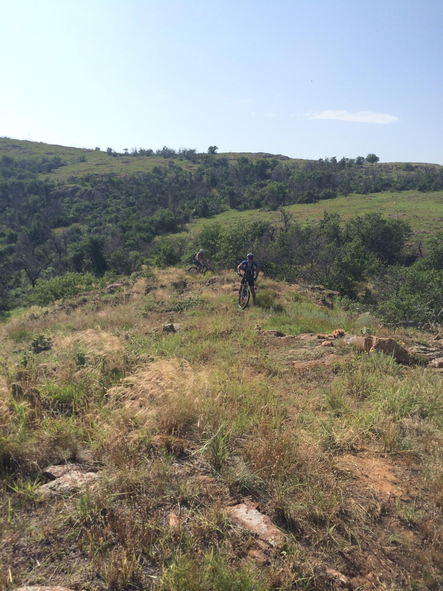 Two mountain bikers navigate a rocky trail on a grassy hillside under a clear blue sky. The landscape features rolling hills and patches of trees, creating a natural outdoor setting for biking. Lake Lawtonka Trails mountain bike trail.
