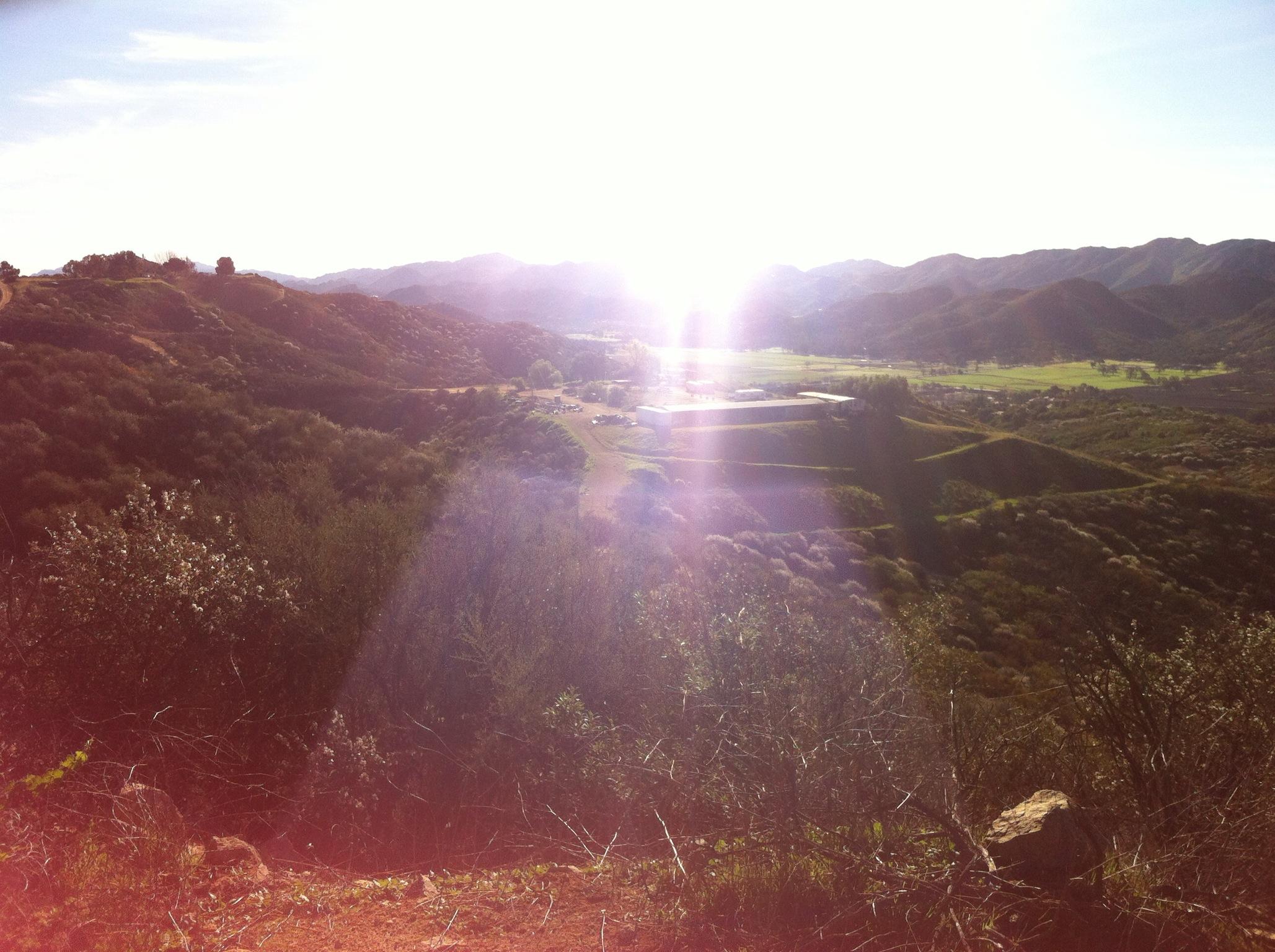A sunlit view of rolling hills and valleys, with lush greenery and a distant building, illuminated by a bright, glowing light. The image captures the natural landscape under a blue sky, showcasing the beauty of nature. Los Robles Trail (Western Section) mountain bike trail.