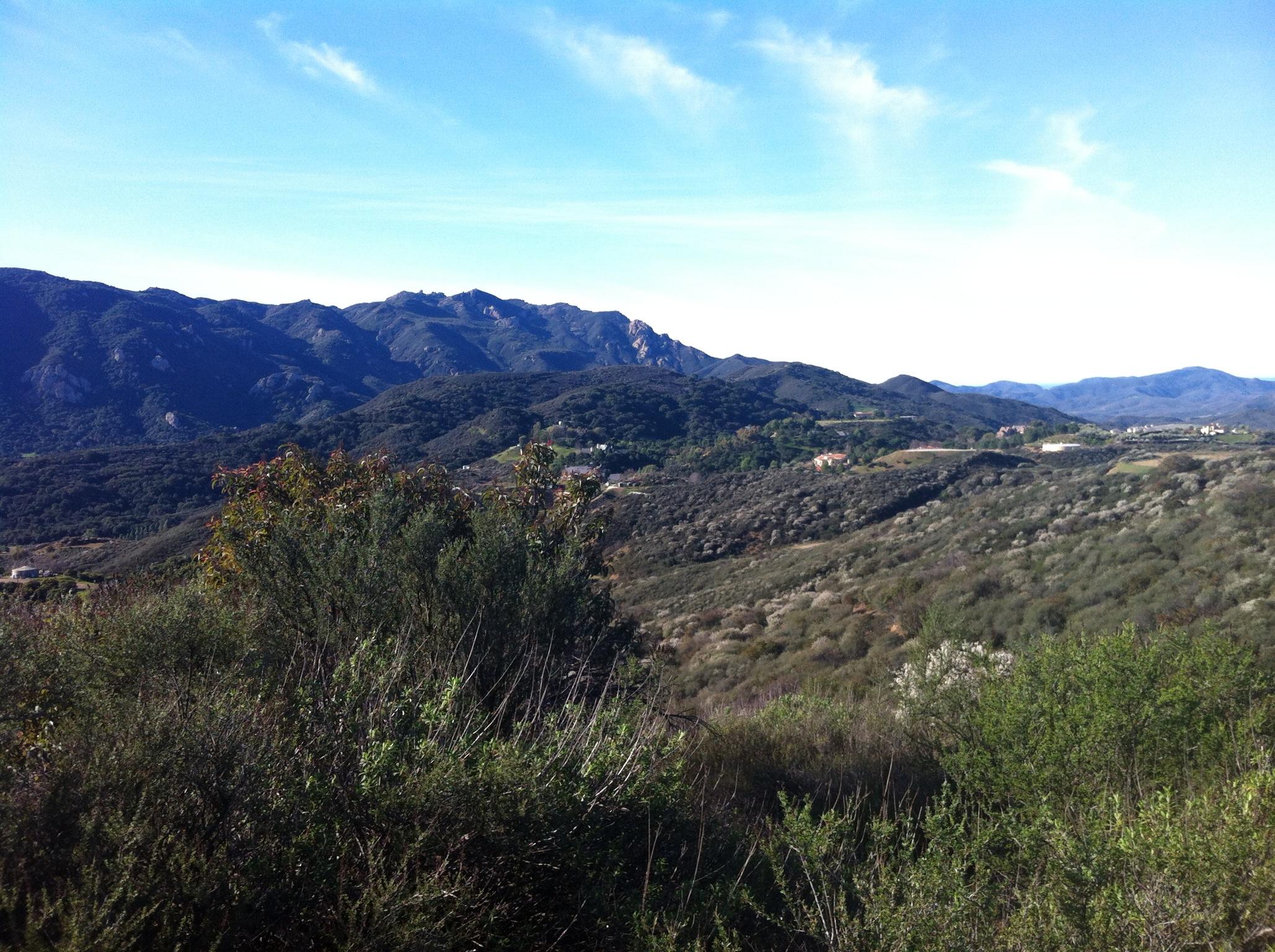 A scenic view of rolling hills and mountains under a clear blue sky, featuring lush greenery and shrubs in the foreground. The landscape showcases varying elevations, with distant mountains providing a natural backdrop. Los Robles Trail (Western Section) mountain bike trail.