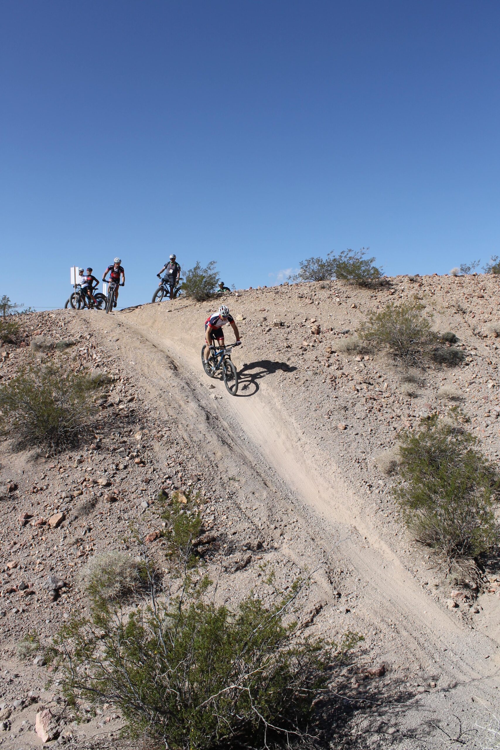 A mountain biker descends a sandy slope in a rugged, arid landscape, with several other cyclists standing on a higher terrain in the background. Clear blue skies and sparse vegetation complete the scene. Bootleg Canyon mountain bike trail.