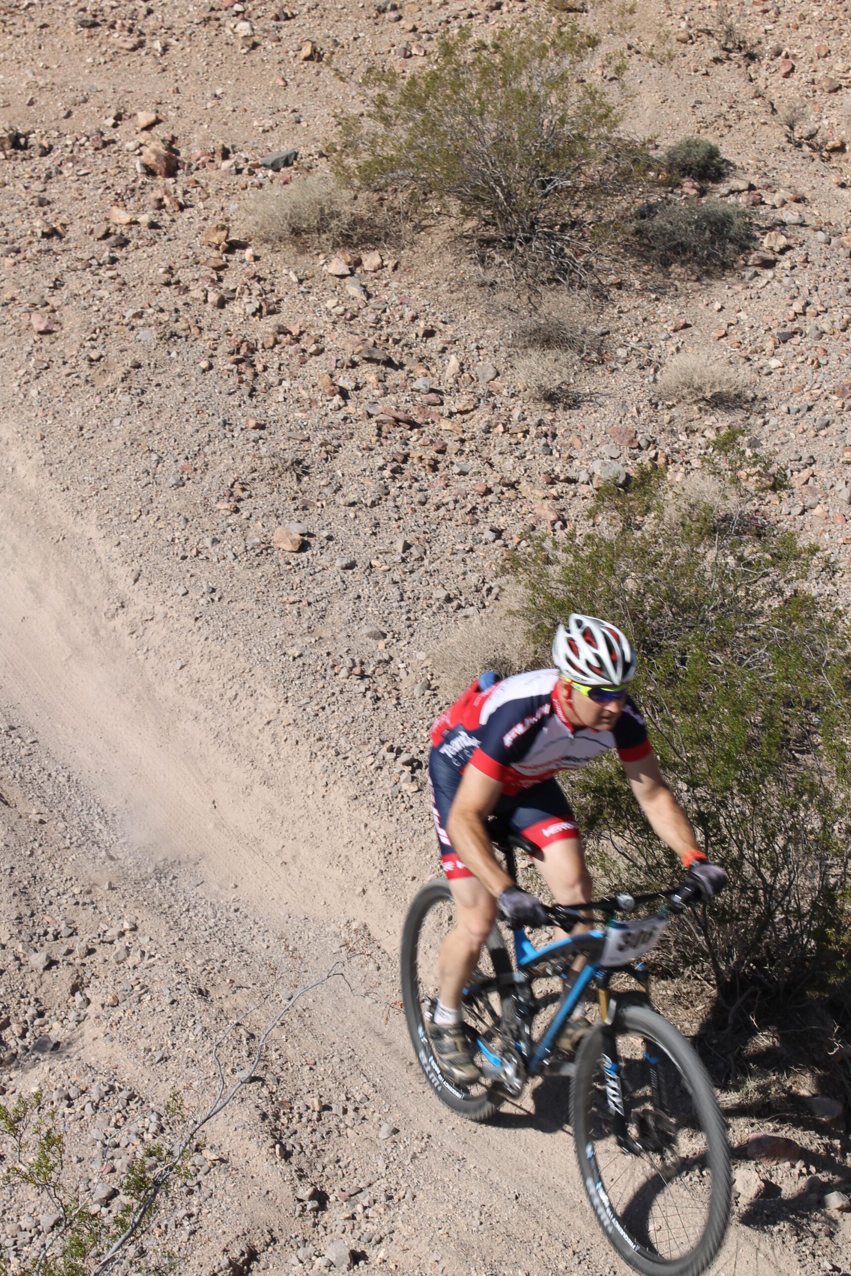 A mountain biker riding on a rugged dirt trail through a rocky landscape. The cyclist is wearing a red and white jersey, black shorts, and a helmet, and is leaning forward on a blue mountain bike. Sparse vegetation and small rocks are visible in the background. Bootleg Canyon mountain bike trail.