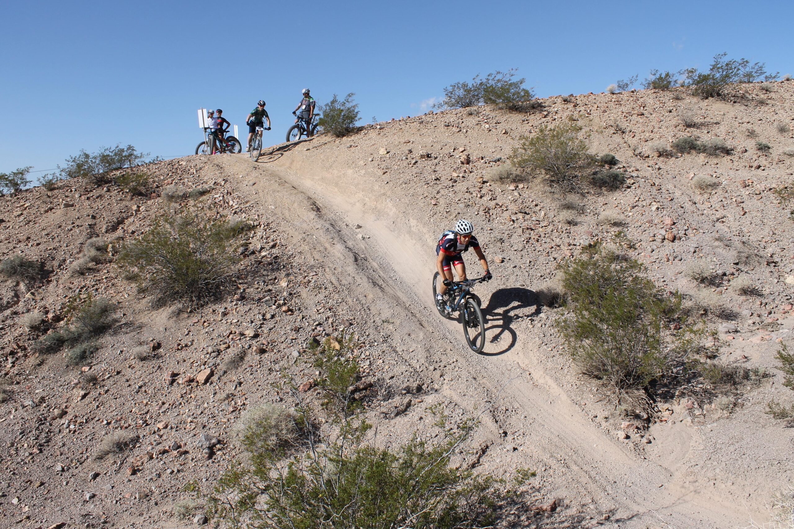A mountain biker navigates a rocky, dirt trail while three other cyclists observe from the top of a hill under a clear blue sky. Sagebrush and scattered rocks are visible on the hillside. Bootleg Canyon mountain bike trail.