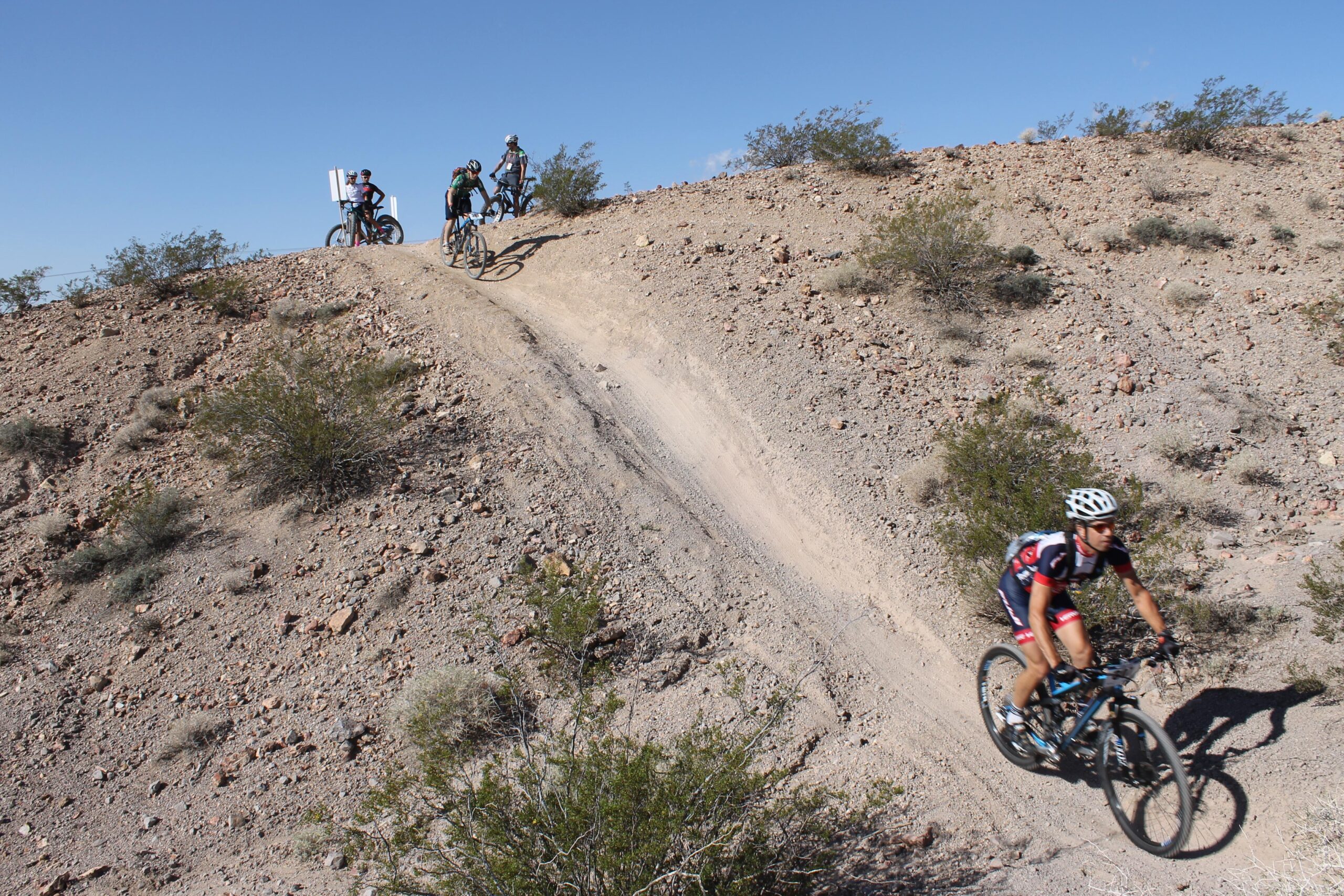 A mountain biker descends a dirt trail on a rugged terrain, surrounded by sparse vegetation under a clear blue sky. In the background, two other cyclists are seen at the top of the hill, preparing to ride down the slope. The scene captures the excitement and challenge of off-road cycling. Bootleg Canyon mountain bike trail.