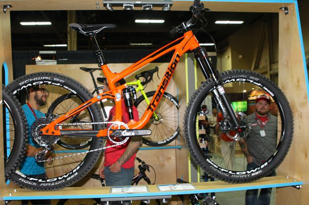 Transition Patrol: Alt text: A bright orange mountain bike displayed on a wooden stand at an exhibition, showcasing its large tires and suspension. Two people are visible in the background, engaging with the display.