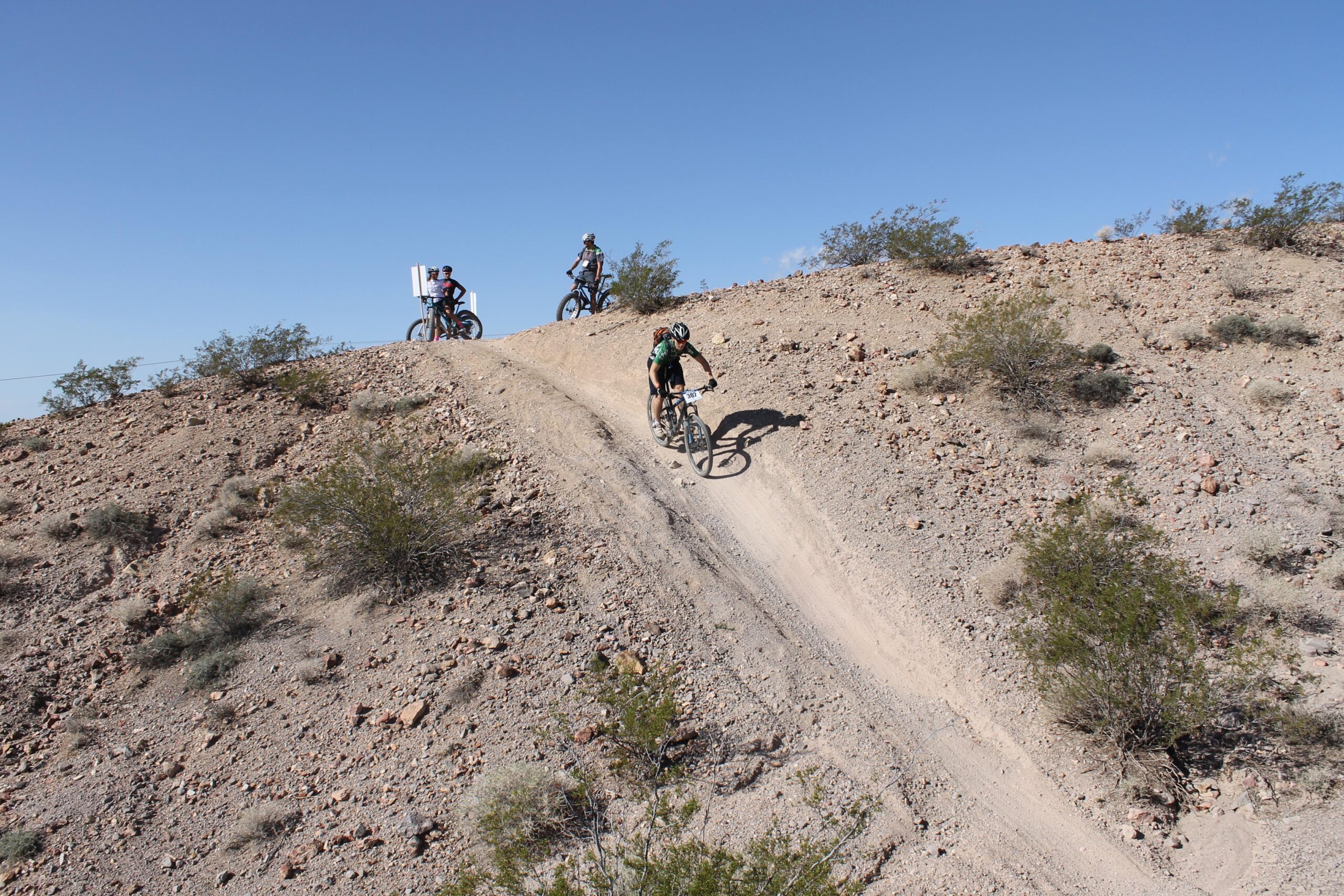 A mountain biker navigates a downhill dirt trail surrounded by rocky terrain and sparse shrubs, while two other bikers are positioned at the top of the hill. The scene is set under a clear blue sky. Bootleg Canyon mountain bike trail.