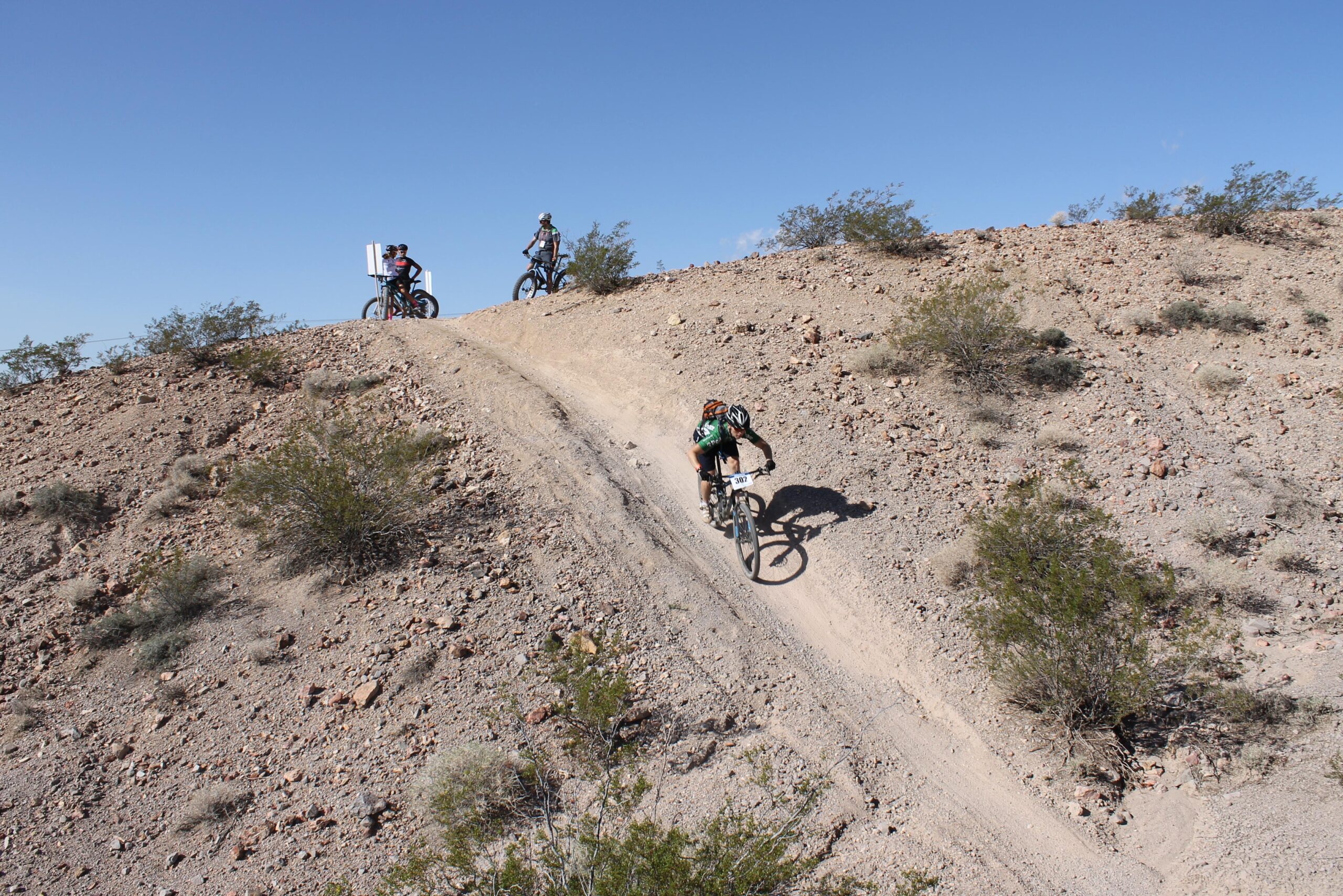 Three mountain bikers are riding on a dirt trail in a desert landscape. One rider is descending a steep section, while two others are positioned at the top of the hill, preparing to descend. The terrain is rugged, with sparse shrubs and rocky ground under a clear blue sky. Bootleg Canyon mountain bike trail.