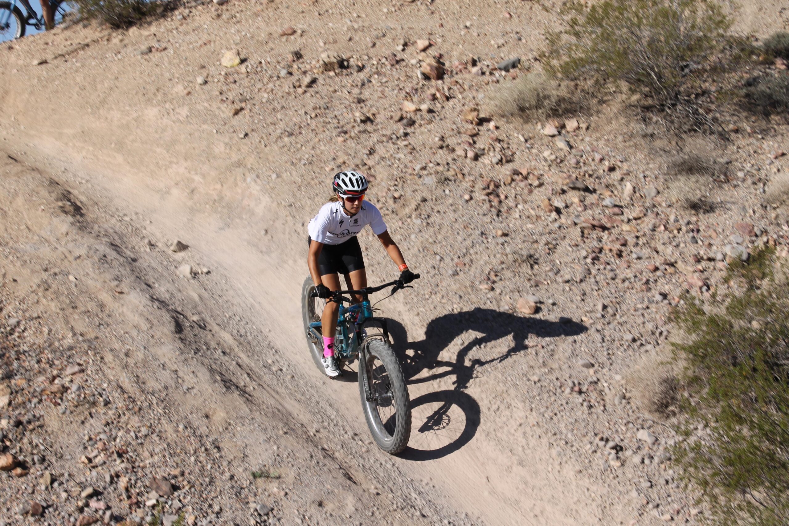 A person riding a mountain bike on a dusty, rocky trail surrounded by sparse vegetation and rocks. The cyclist is wearing a helmet, a white shirt, and black shorts, with pink socks visible. The terrain is hilly, and the sun is shining, casting a shadow of the rider on the ground. Bootleg Canyon mountain bike trail.