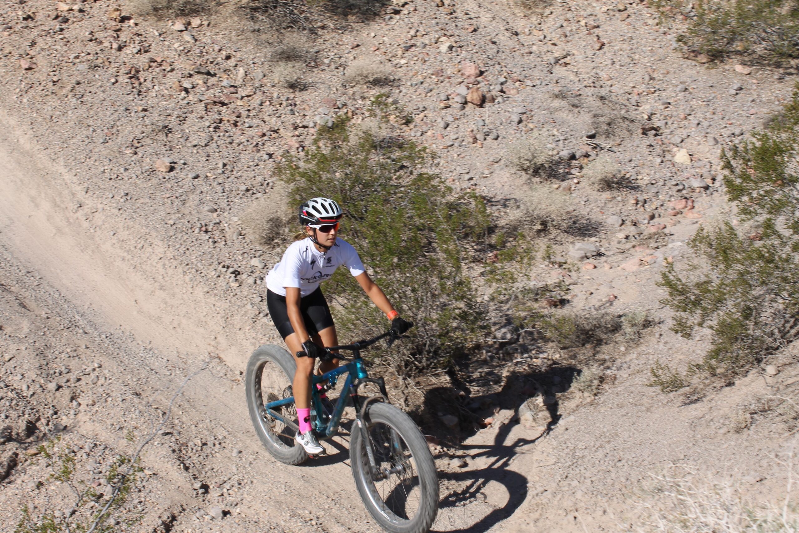 A person riding a mountain bike on a dirt trail surrounded by rocky terrain and shrubs. The cyclist is wearing a white jersey, black shorts, and a helmet, with bright pink socks visible. Bootleg Canyon mountain bike trail.