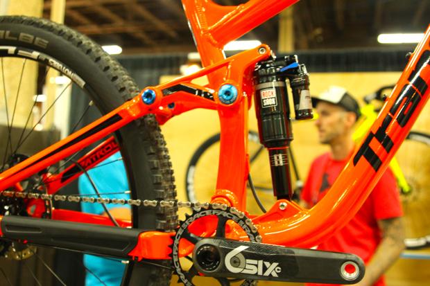 Transition Patrol: Close-up view of a vibrant orange mountain bike frame, highlighting its suspension system and drivetrain. In the background, a person in a red shirt and a cap is partially visible, along with other bikes. The focus is on the bike's components, showcasing the craftsmanship and design details.