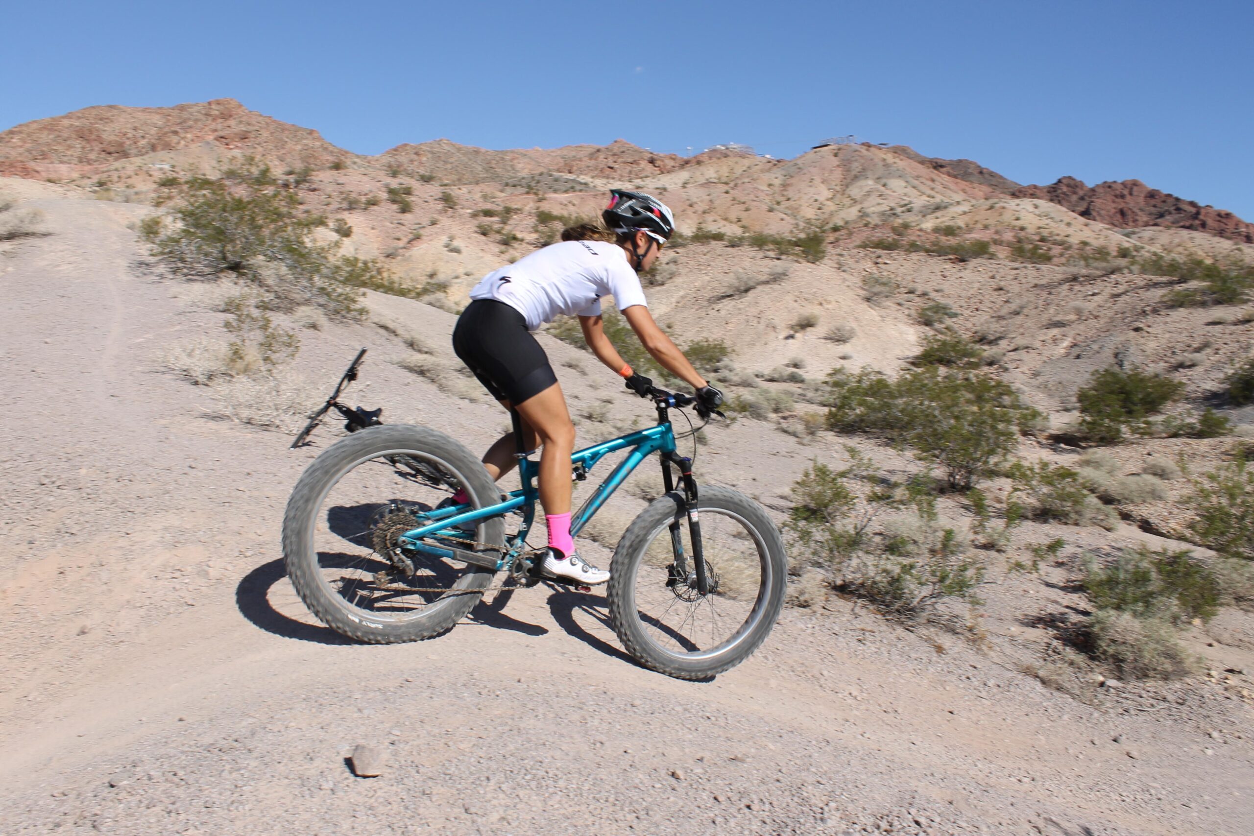 A person riding a turquoise mountain bike on a dirt trail in a rocky desert landscape, with mountains in the background and clear blue skies. The cyclist is wearing a white shirt, black shorts, and pink socks. Bootleg Canyon mountain bike trail.