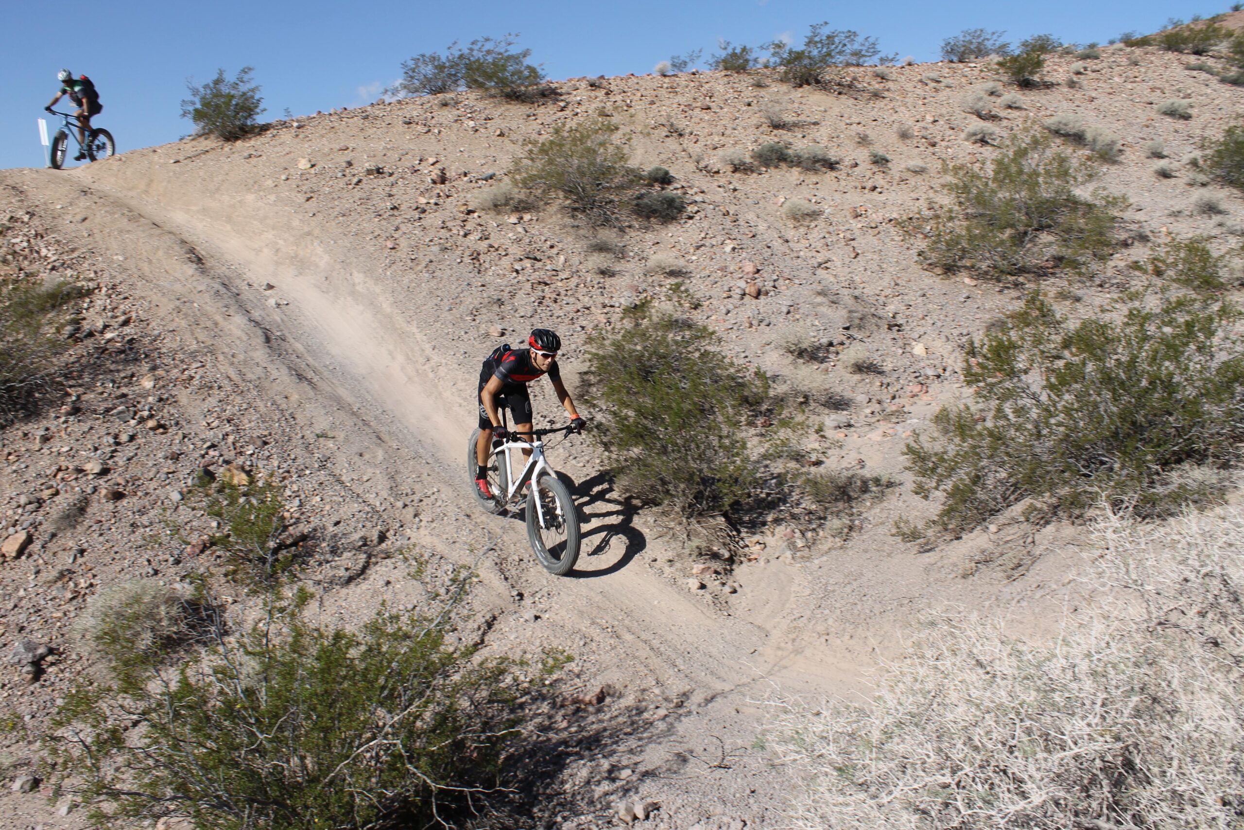 Two mountain bikers navigate a rocky trail on a hillside. One rider, dressed in black with red accents, is leaning forward as they descend a slope on a white bike, while the second rider in the background approaches on a dark-colored bike. The landscape features dry, barren terrain with sparse vegetation and rocky ground under a clear blue sky. Bootleg Canyon mountain bike trail.