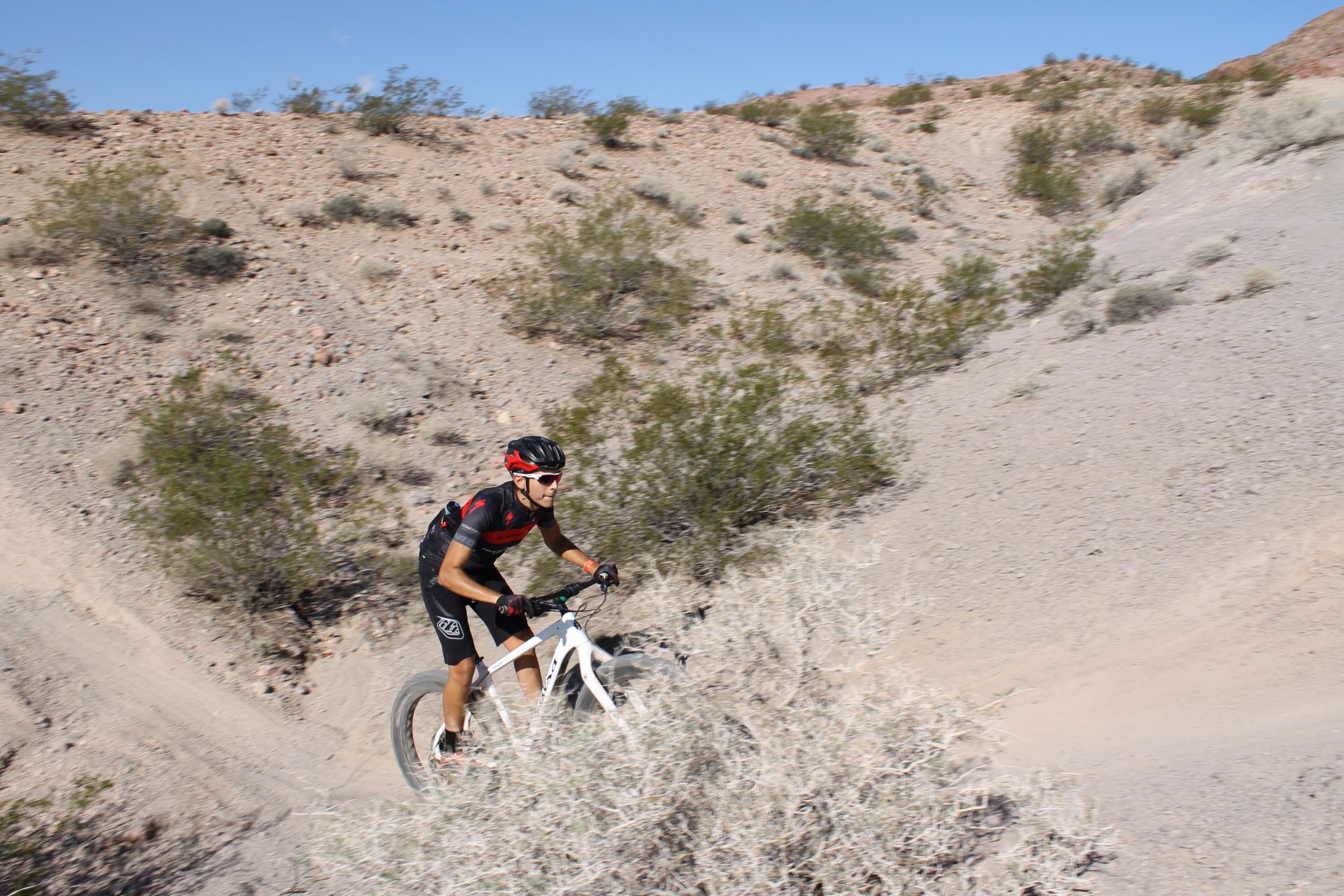 A cyclist riding a mountain bike along a dirt trail in a rocky, arid landscape with sparse vegetation and a clear blue sky. Bootleg Canyon mountain bike trail.
